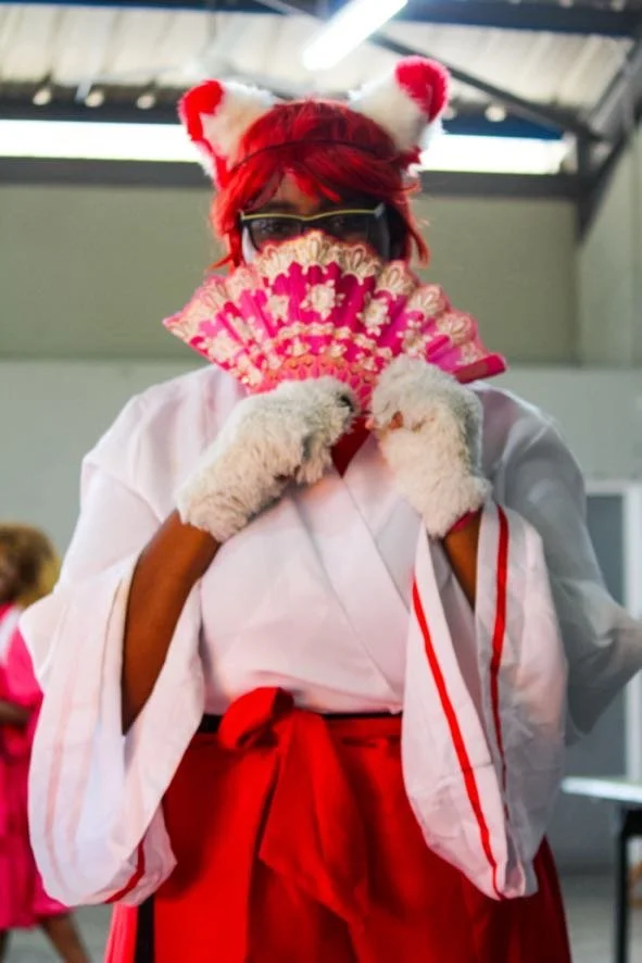 Person wearing a kawasaki karate gi with red belt, fur gloves, red hair, and a headband with cat ears, holding a pink floral fan in front of her face.