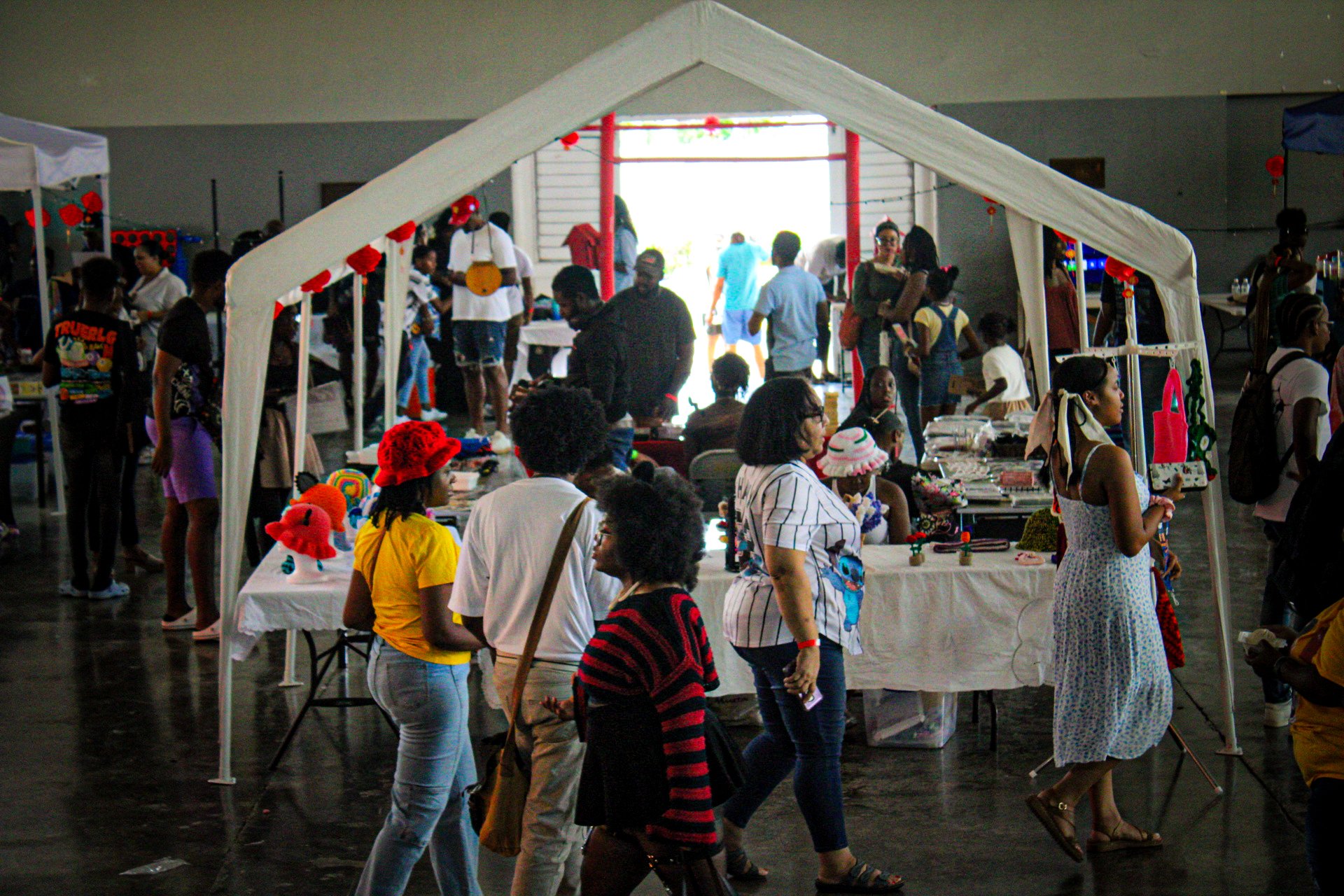 People shopping and browsing at a craft fair inside a large building with booths displaying handmade items, including hats and decorations, with some people looking at the items and others walking around or browsing.