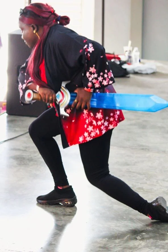 A woman with red hair and wearing a floral kimono-themed robe is kneeling on one knee while holding a large blue and silver sword in a room with a gray floor.