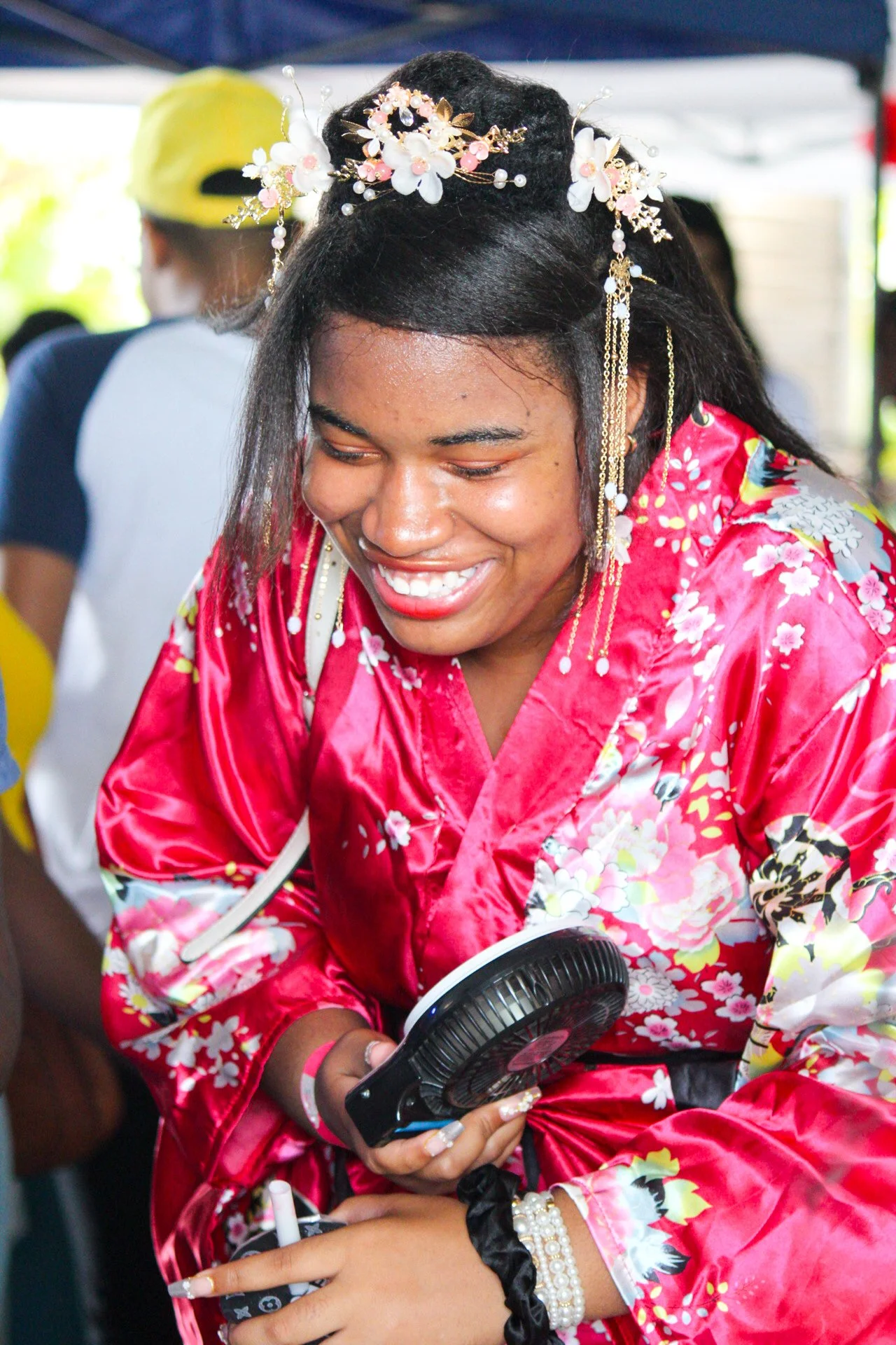 Young woman wearing a red floral kimono and floral hair accessories, smiling while holding a small fan at an outdoor event.