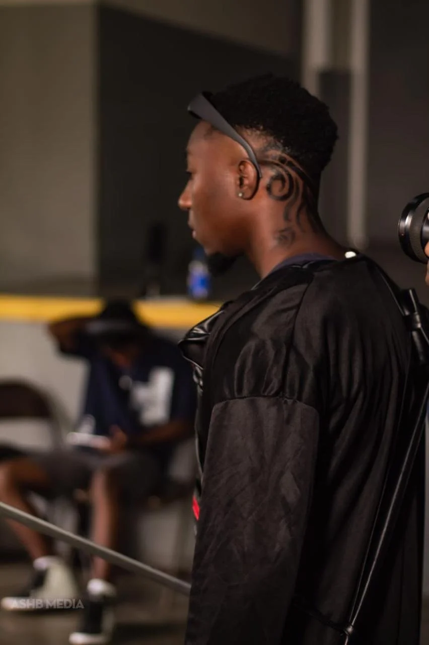 A young man with styled hair and earrings, wearing a black jacket, standing with his eyes closed in a room with blurred background, including another person sitting and using a phone.