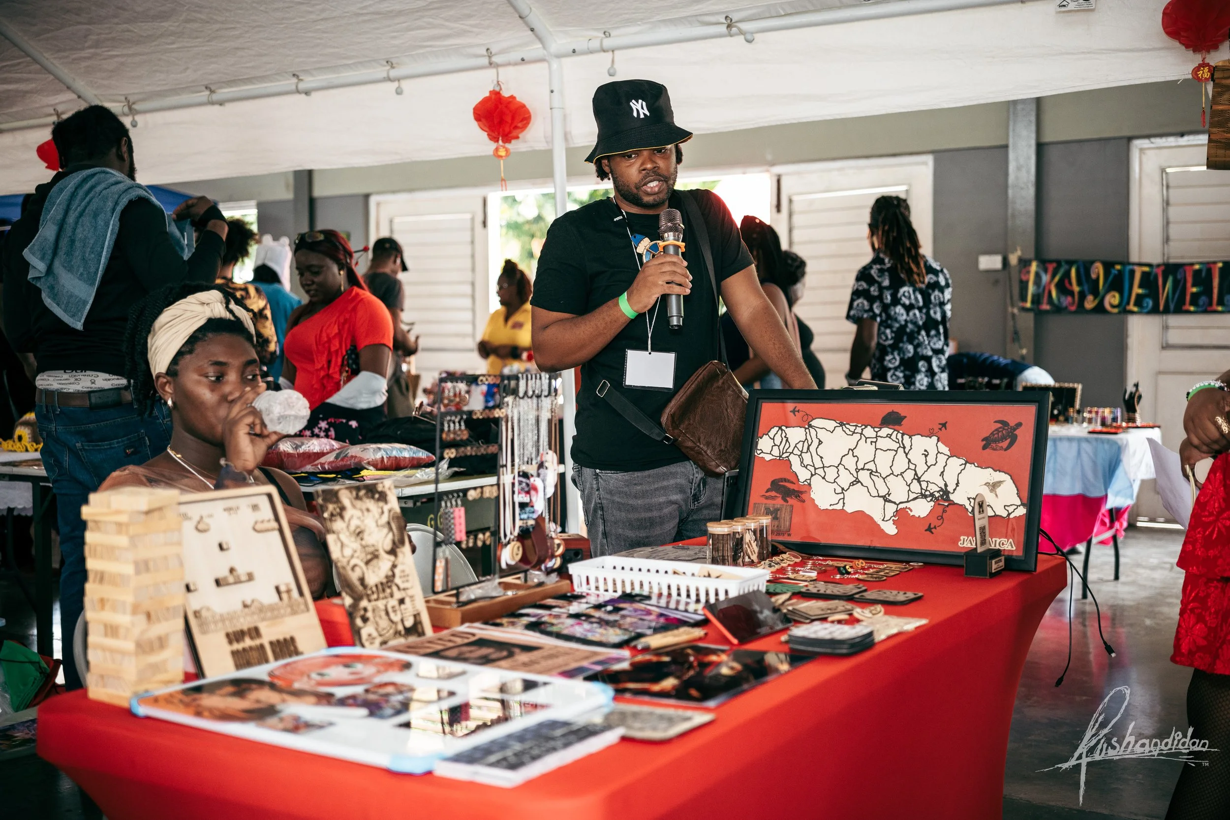 A man in a black t-shirt and bucket hat is speaking into a microphone at a booth with jewelry and a map of Jamaica on display, with several people browsing and shopping around him.