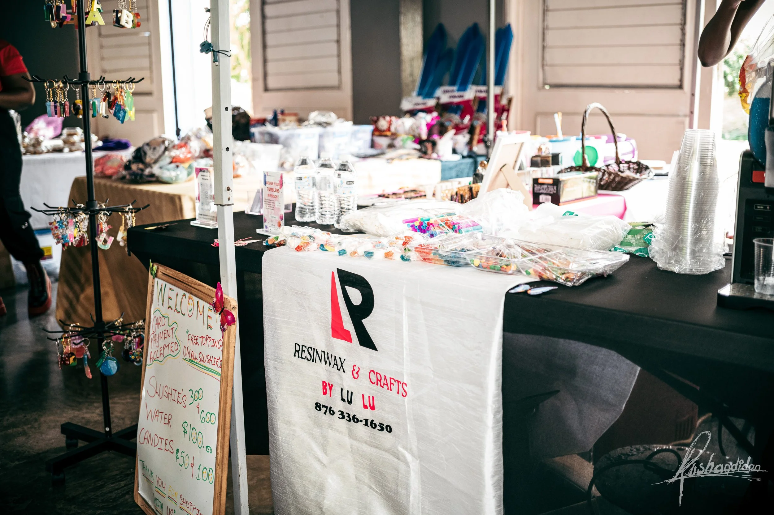 Indoor craft market with tables displaying jewelry, water bottles, candies, and other handmade items. Signboard advertises various snacks and prices. The main table has a white sign with garage sale details for Resinwax & Crafts by Lu Lu. Sunlit back