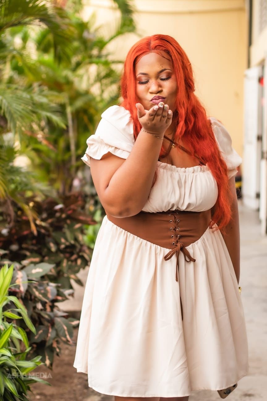 A woman with long red hair wearing a beige dress with a brown corset blowing a kiss outdoors.