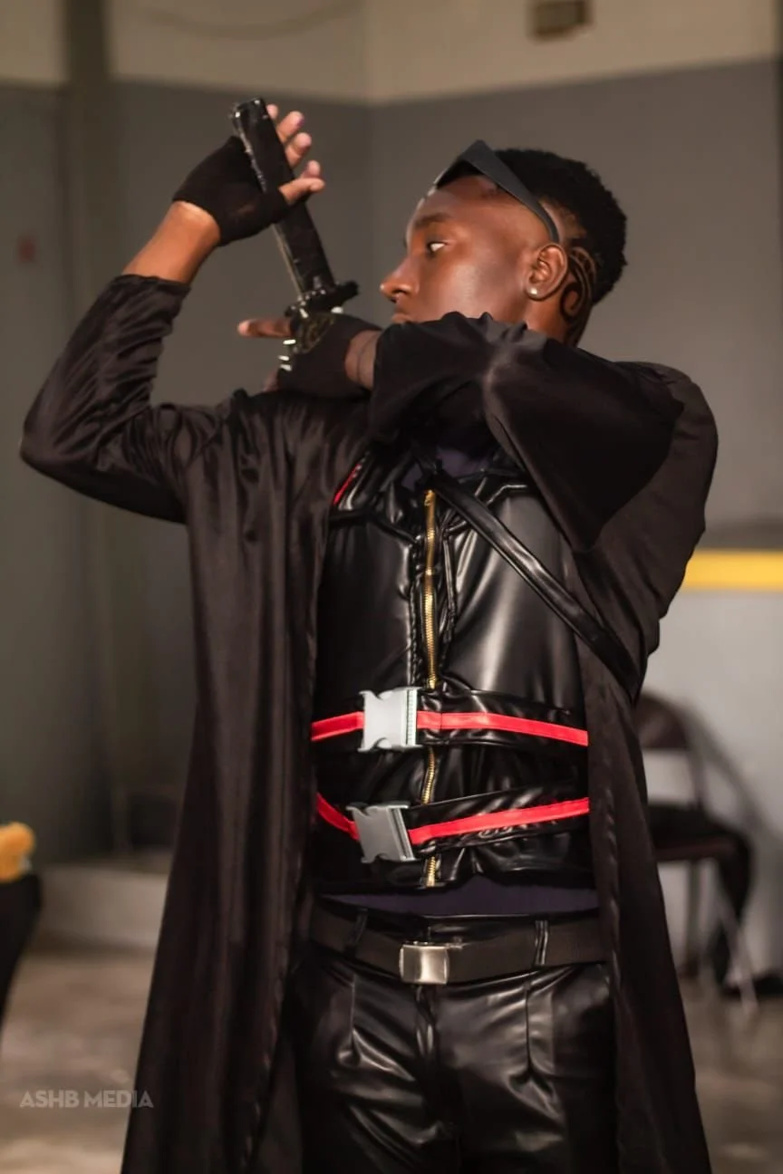 A person dressed in black, wearing a futuristic vest with red accents, is holding a gun to their head in an indoor setting.