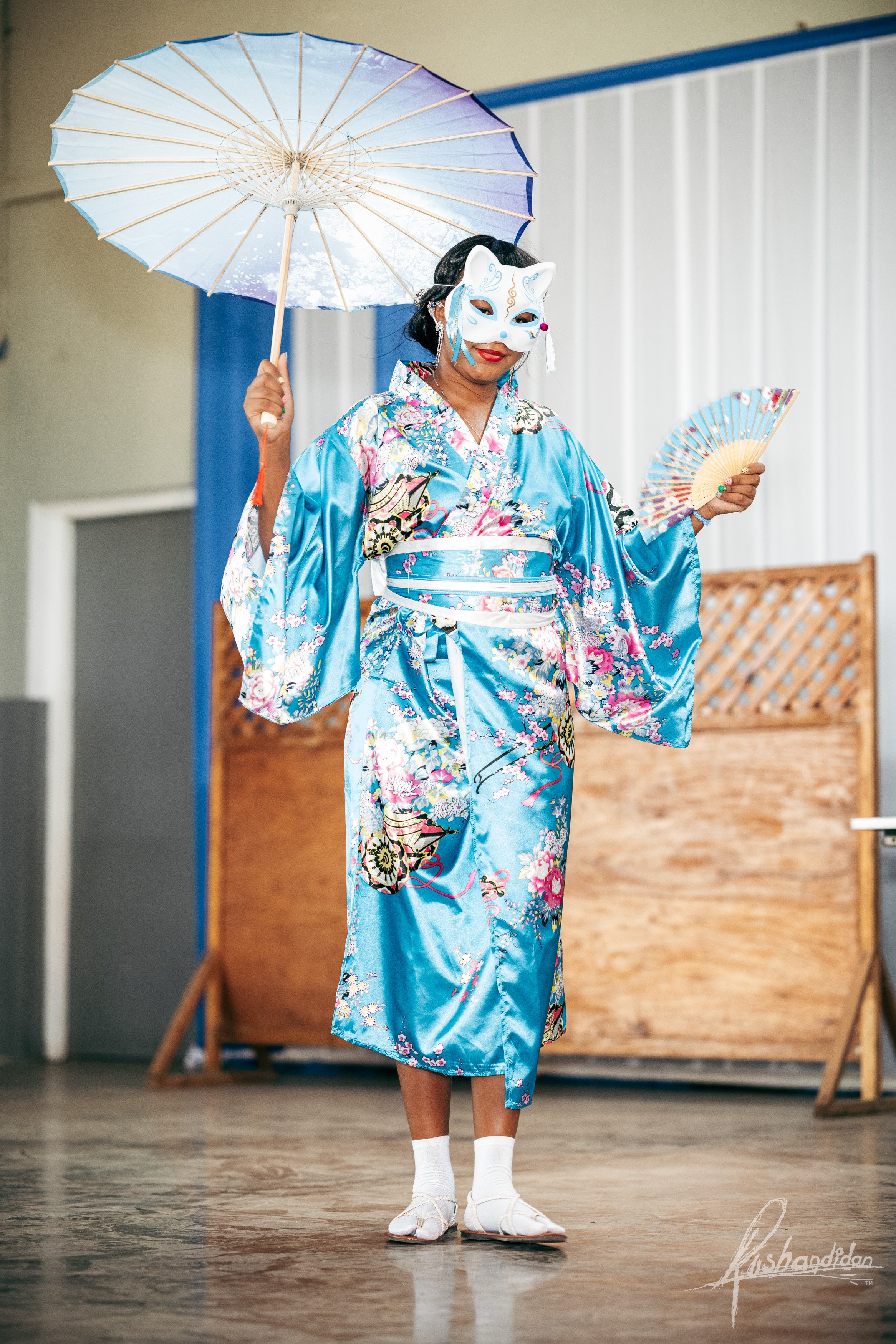 A performer dressed in a vibrant blue kimono decorated with floral and traditional patterns, holding a paper fan in one hand and an umbrella in the other, wearing a white and blue fox mask, standing on a stage with a wooden backdrop.