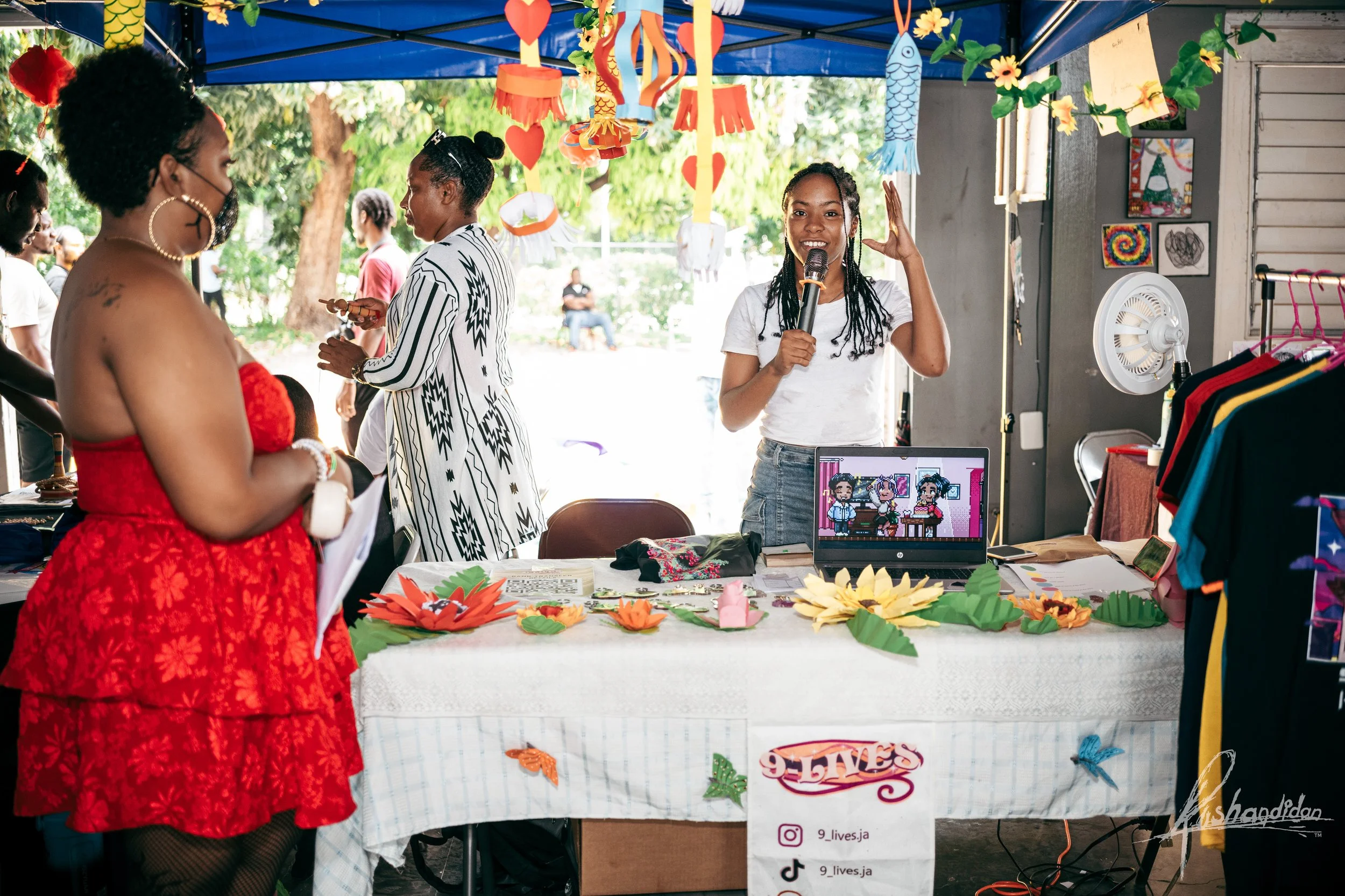 A woman with long braided hair speaking into a microphone at an outdoor event booth decorated with paper flowers and hanging paper lanterns, with several people around.