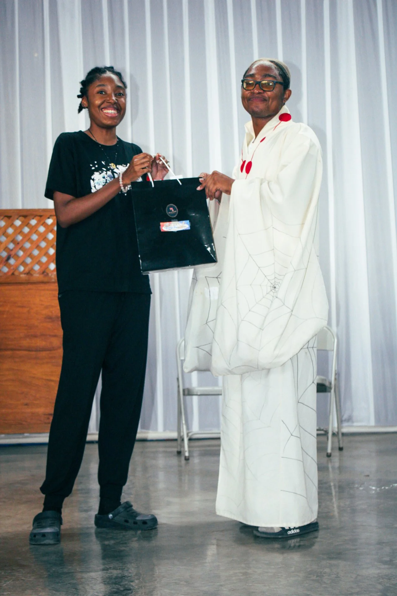 A woman wearing a black t-shirt and black pants is smiling and handing a black gift bag to an older man dressed in a white outfit with spider web designs. They are inside a room with a wooden panel and white curtains in the background.