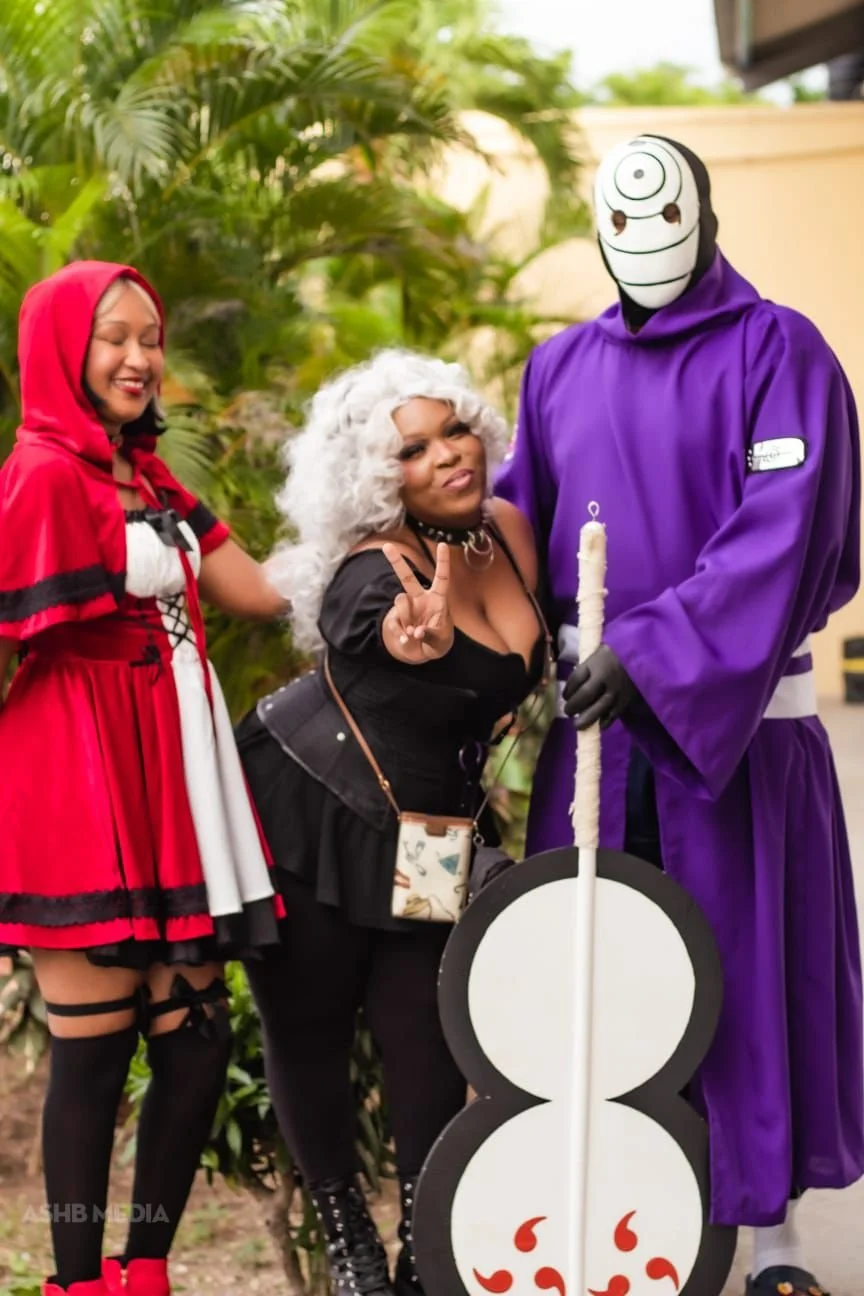 Three people in costumes posing outdoors. One person is dressed as Little Red Riding Hood, another in a black outfit with white curly wig making a peace sign, and the third as a character with a white face mask with black markings, purple robe, and h