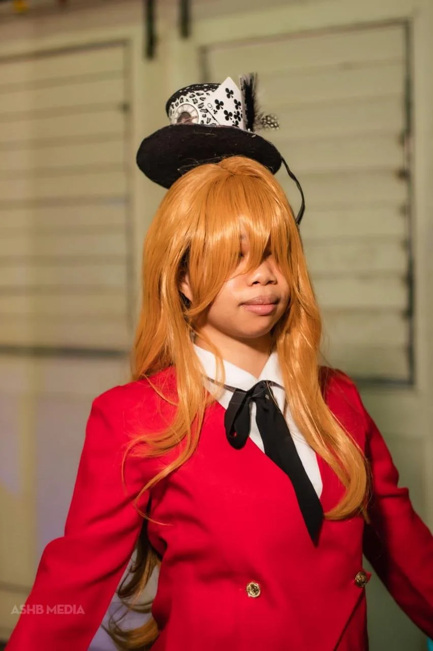 Woman with long orange hair wearing a red blazer with gold buttons, white shirt, and black tie, standing indoors with beige walls and a garage door in the background, balancing a black hat with a card and a small feather on her head.