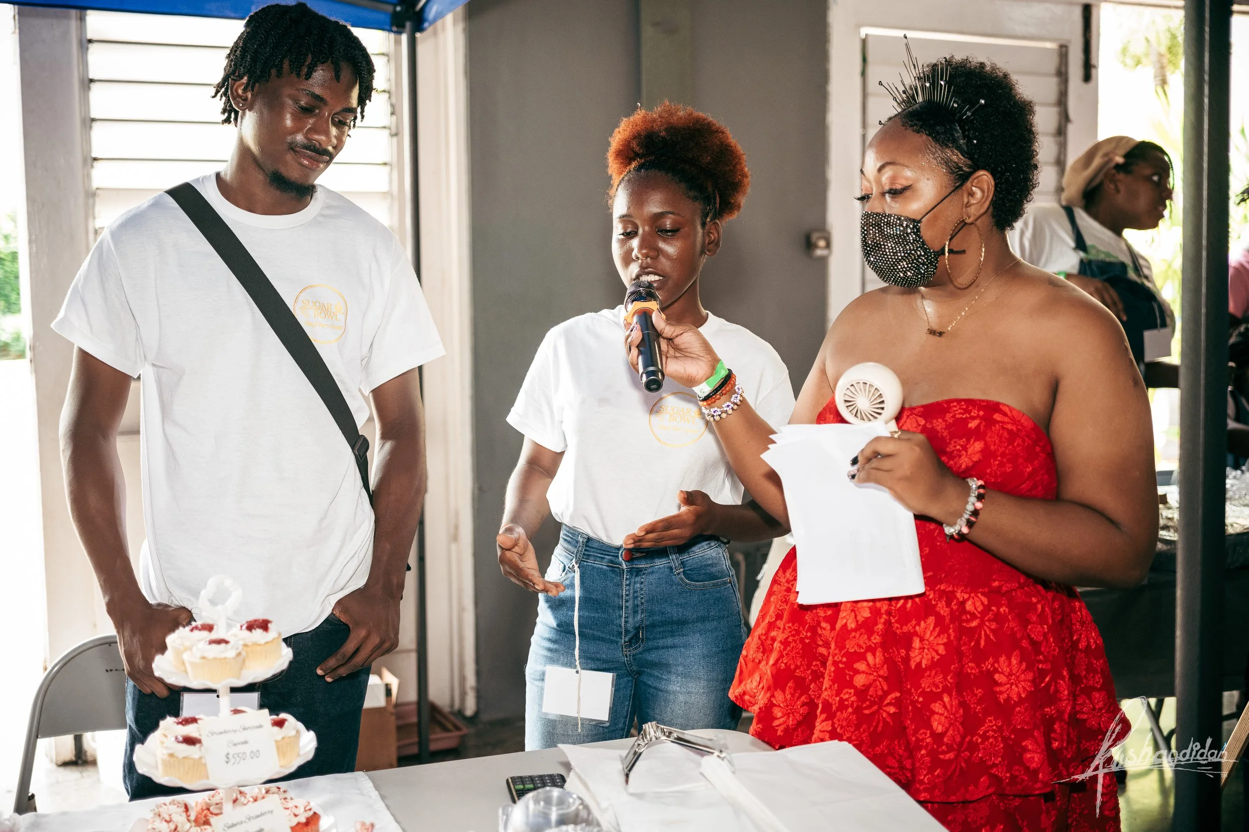 A woman in a red dress and face mask holds a paper and a paper fan, speaking into a microphone held by a woman in a white shirt. A man in a white shirt stands nearby with hands in pockets. They are indoors, surrounded by cakes on a table, with a woma