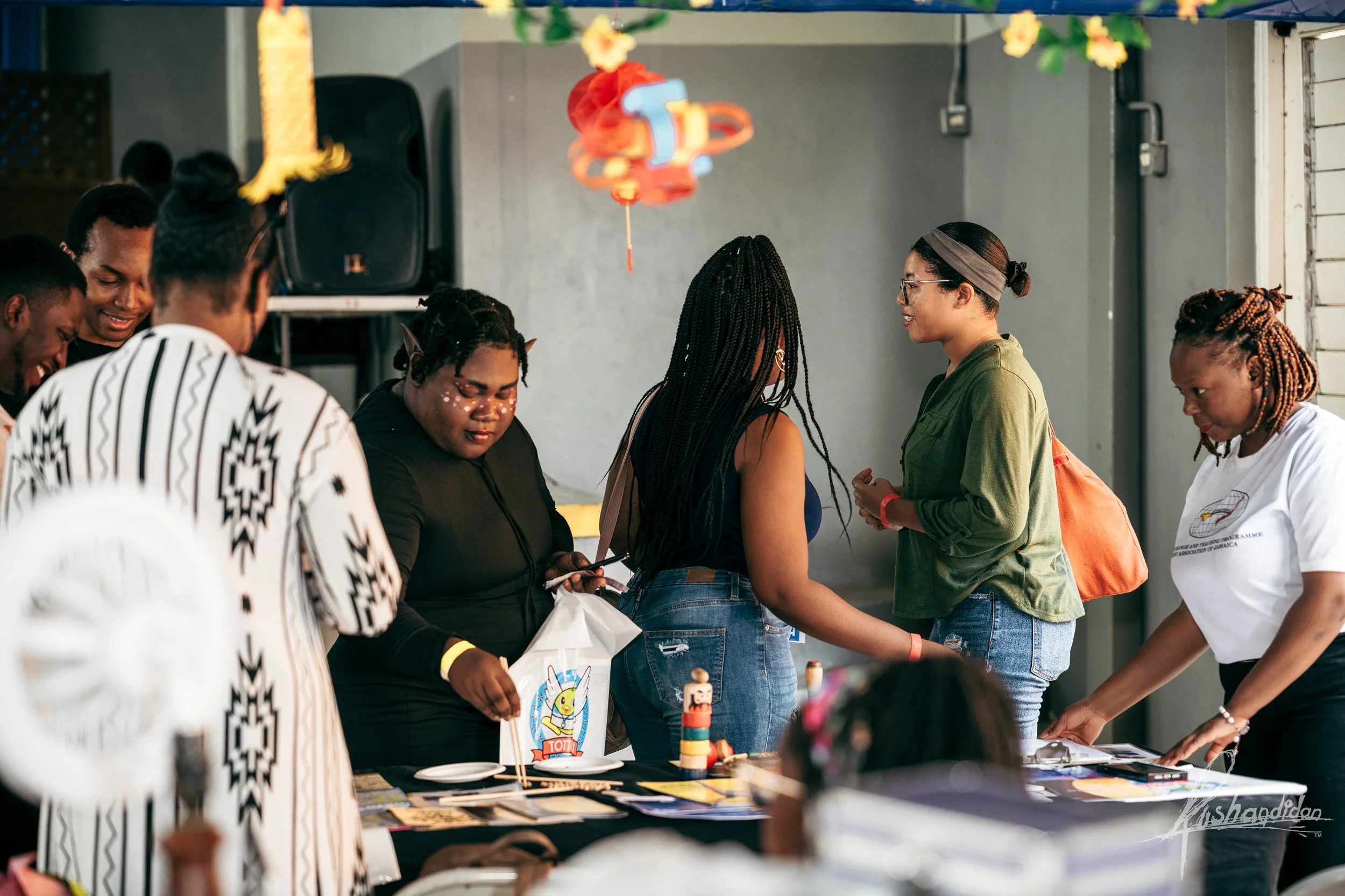 Group of people gathered around a table, engaging in conversation and browsing items at what appears to be an indoor event or fair.