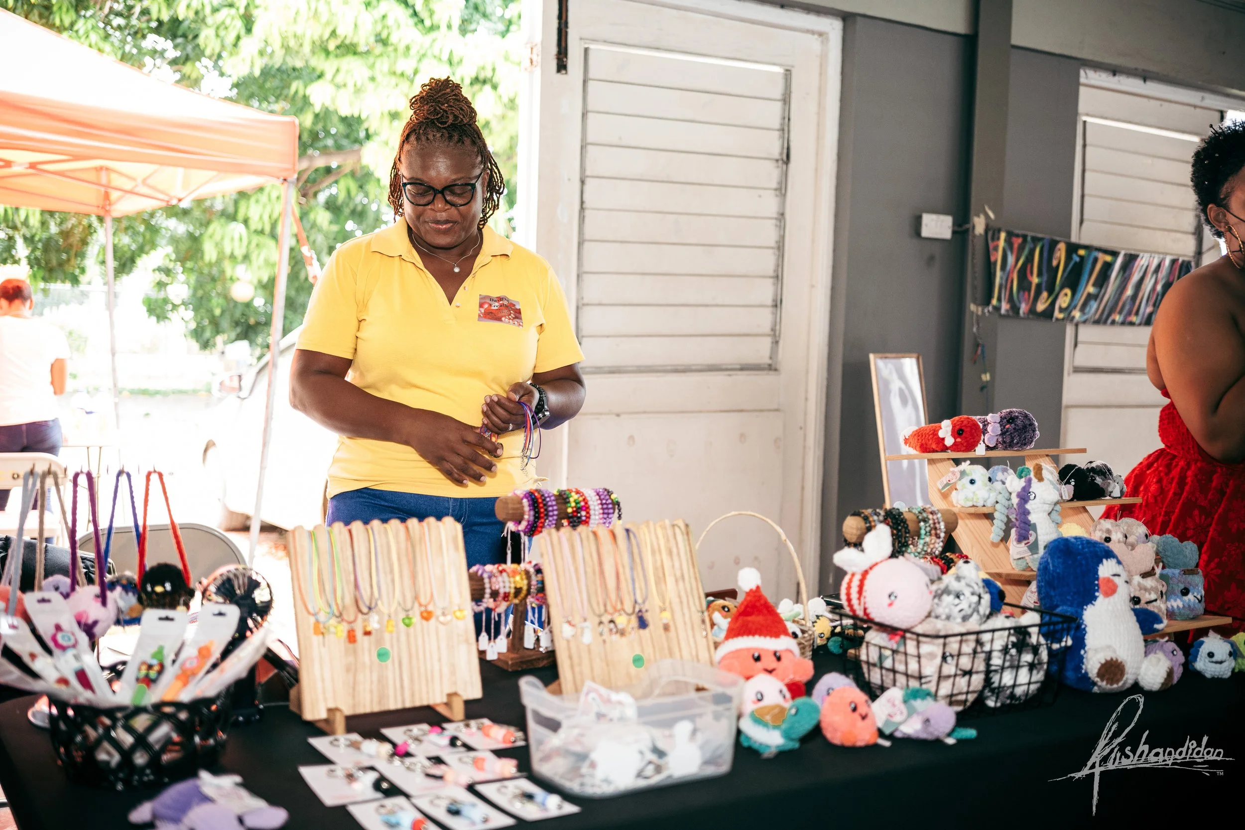 A woman in a yellow shirt browses handmade jewelry at an outdoor craft market stall. The table displays colorful necklaces, earrings, and crocheted plush toys. Other vendors and customers are visible in the background.