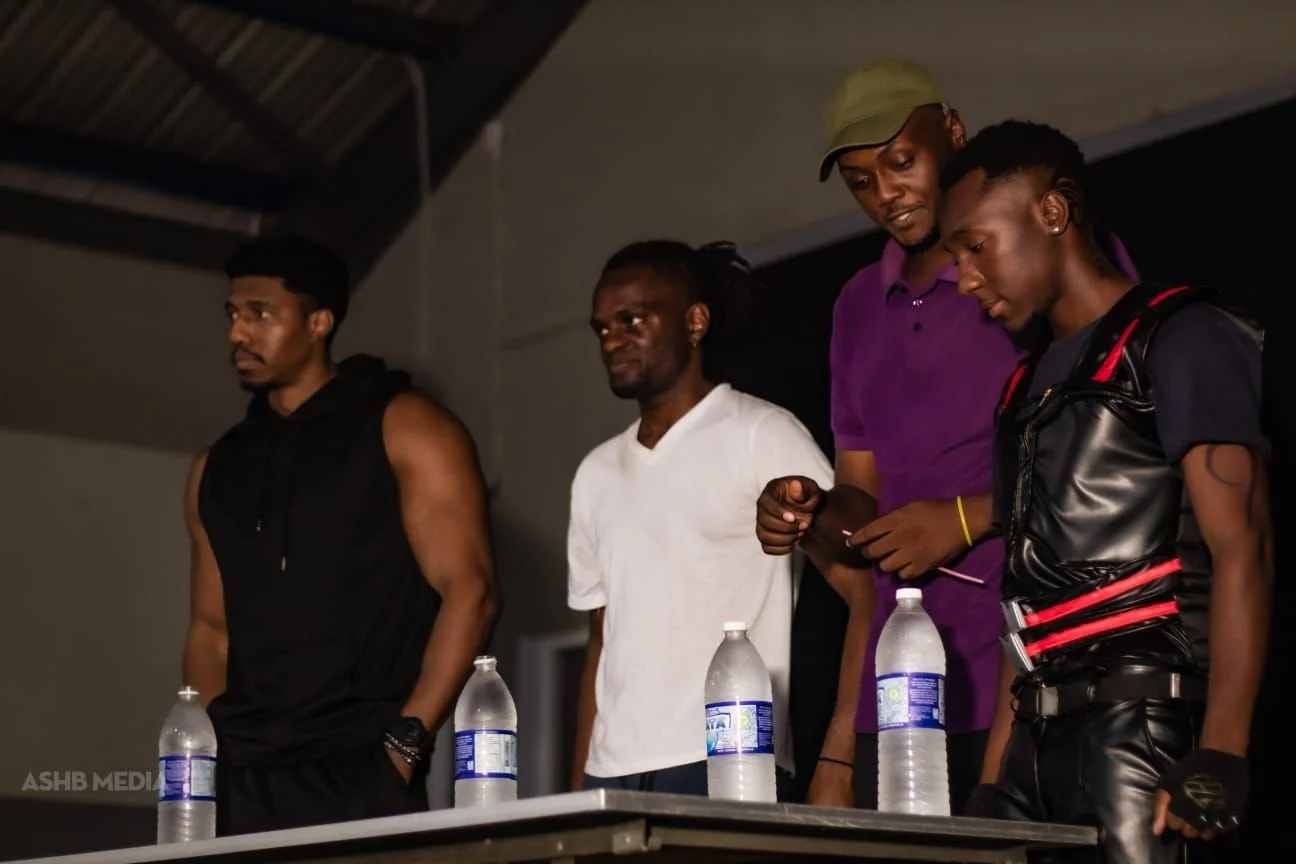 Four individuals are standing behind a table with water bottles. Two are men and two are women, one in a white shirt and others in casual attire. The setting appears to be indoors, possibly at an event or gathering.