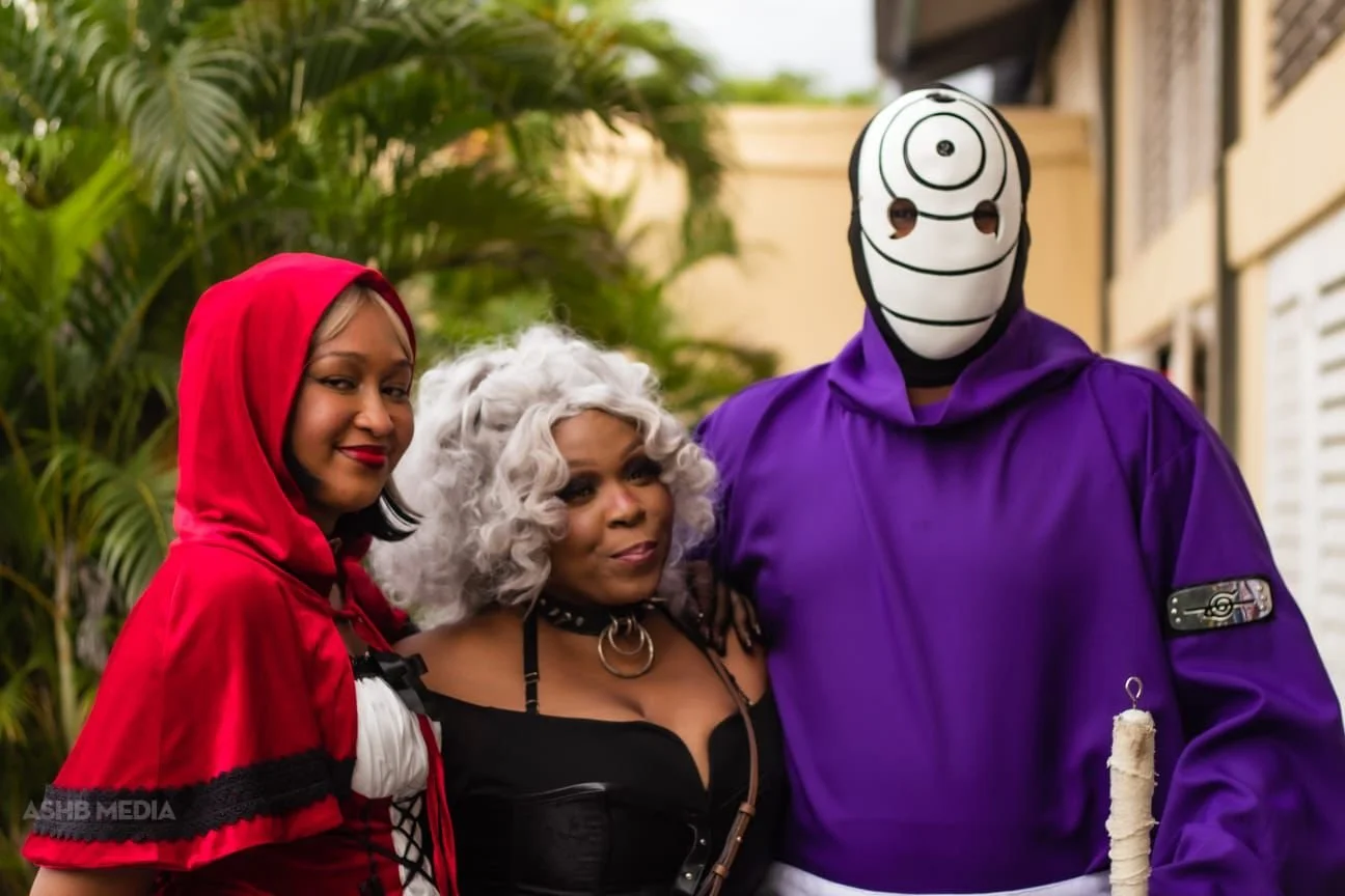 Three people dressed in costumes standing outdoors, with palm trees in the background. One person is dressed as Little Red Riding Hood, another as an older woman with curly gray hair in a black outfit with a choker, and the third is wearing a purple 