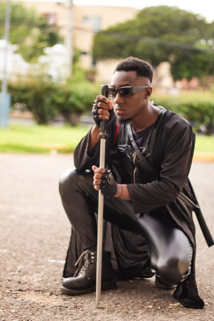A man dressed in black, wearing gloves and sunglasses, kneeling on the ground and holding a crowbar in one hand. He has a serious expression and is posed in an urban outdoor setting.