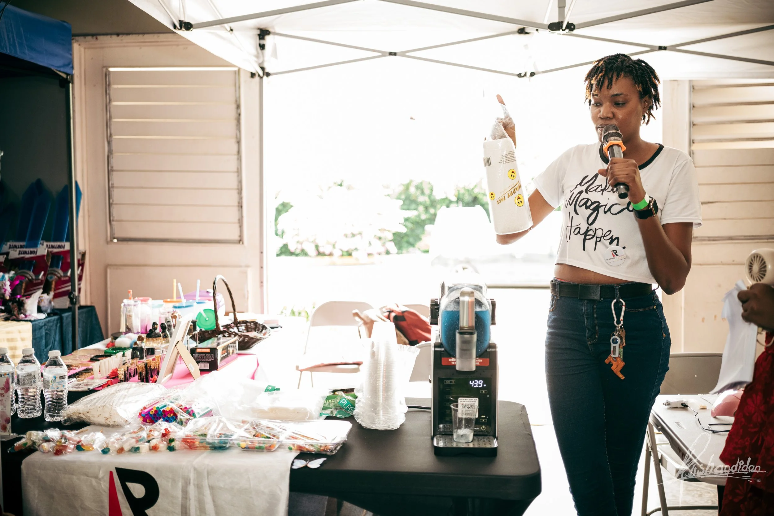 A woman with short curly hair, wearing a white crop top with a phrase, holding a microphone in one hand and talking, standing in a booth with various items on a table, at an outdoor event.