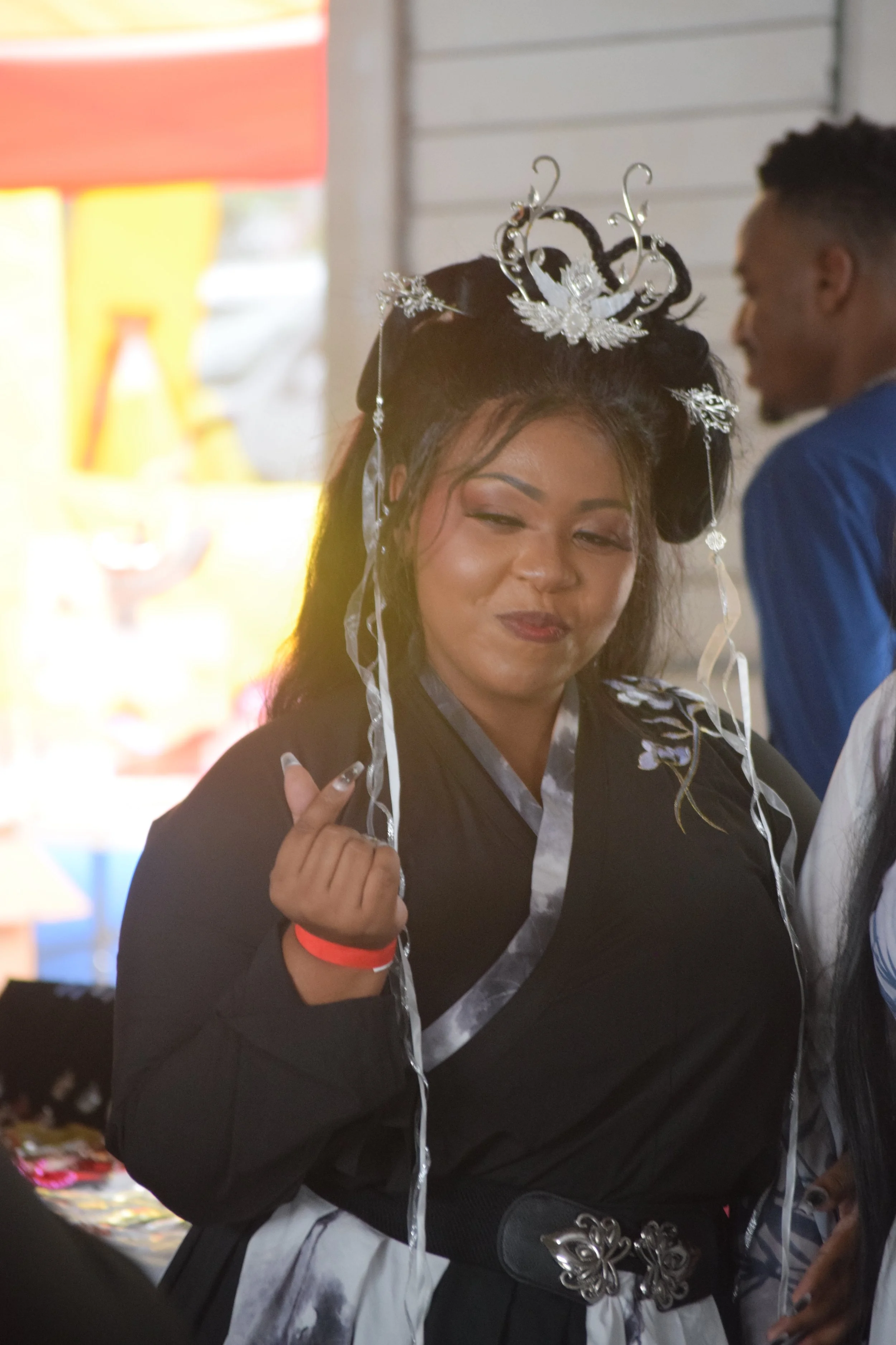 Woman wearing a silver tiara with swirls and wings, smiling, and holding a ribbon, standing indoors at a celebration.