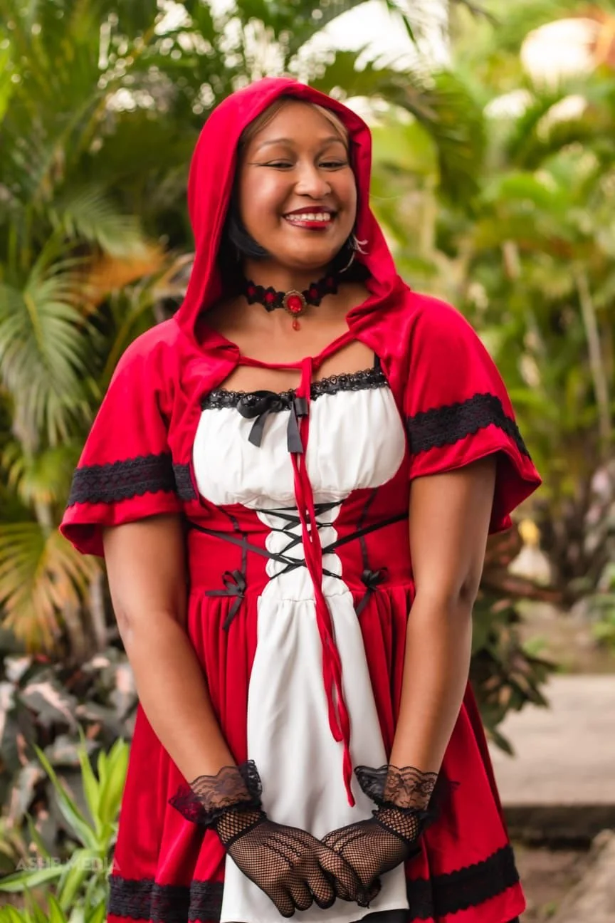 A woman smiling outdoors, dressed in a red hooded cloak with black lace accents, a white dress, lace gloves, and a black choker with a red gemstone. She stands in front of lush green foliage.
