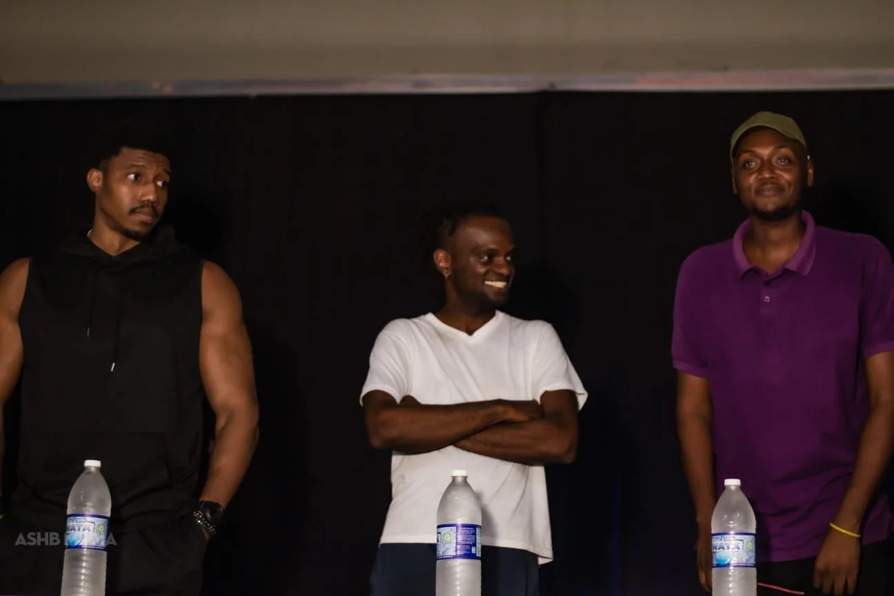 Three men standing side by side in front of a dark background, with bottled water on a table in front of them. The man on the left is wearing a sleeveless black hoodie, the man in the middle is wearing a white T-shirt, and the man on the right is wea