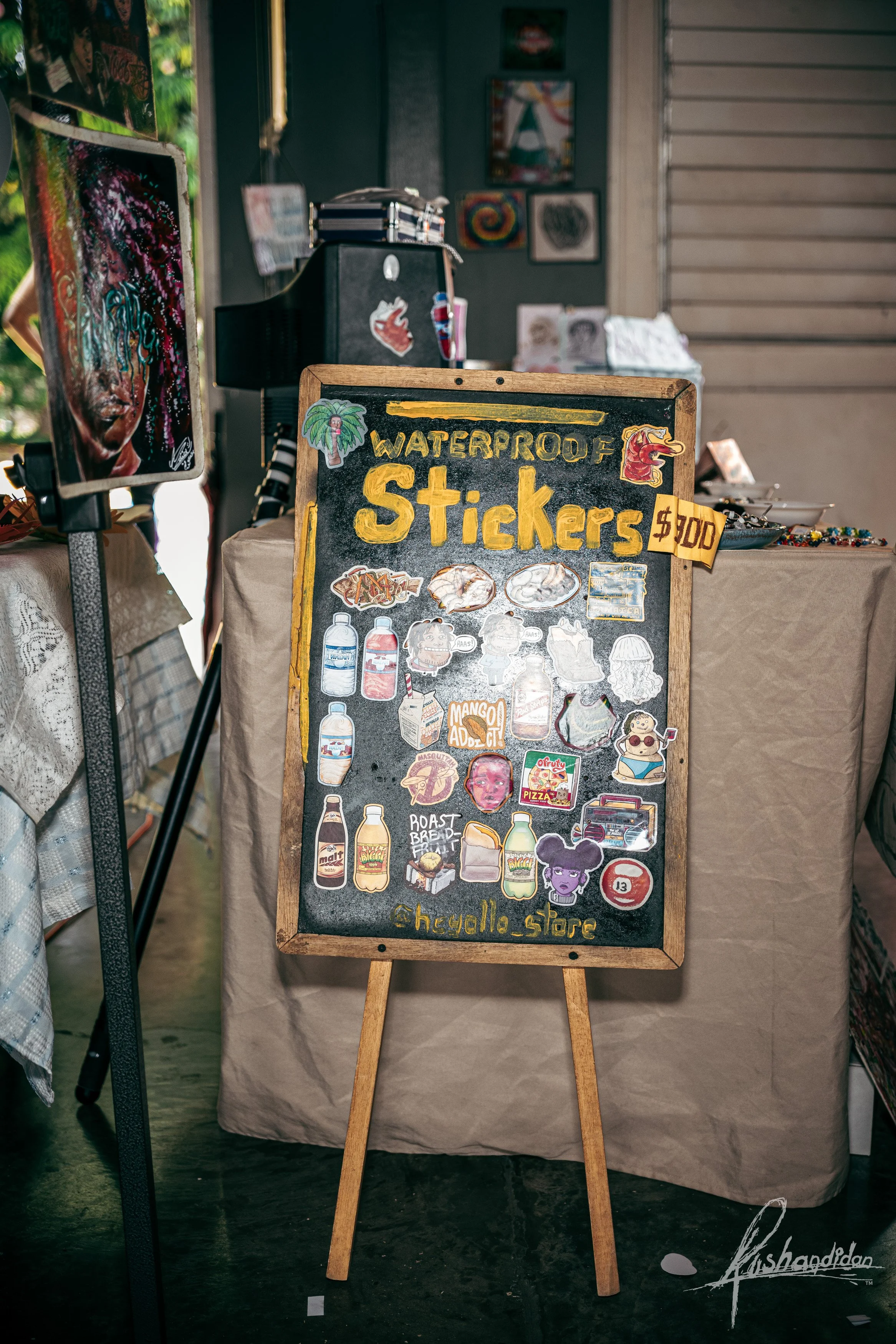 A chalkboard sign advertising waterproof stickers for $1.00 each, decorated with colorful illustrations and stickers, including bottles, food, and characters, with a small sign showing the price, placed inside a shop or market setting.