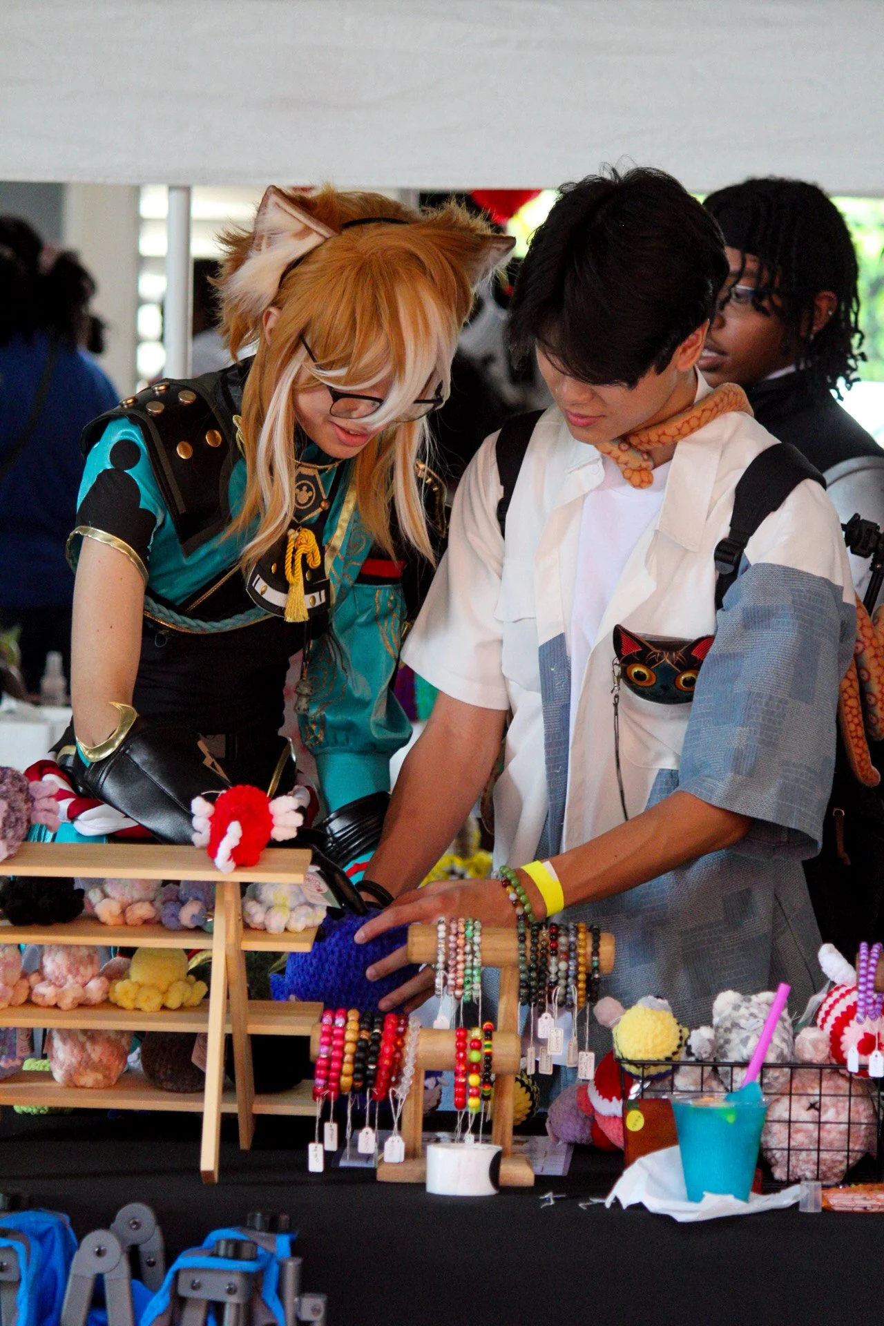 Two people at a craft fair looking at handmade yarn and beaded jewelry displays, surrounded by colorful accessories and decorations.