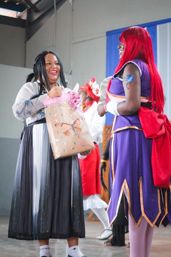 Two women dressed in costumes exchanging gifts, one with long black hair and a striped skirt, and the other in a purple and red costume with long red hair, smiling and holding a gift bag