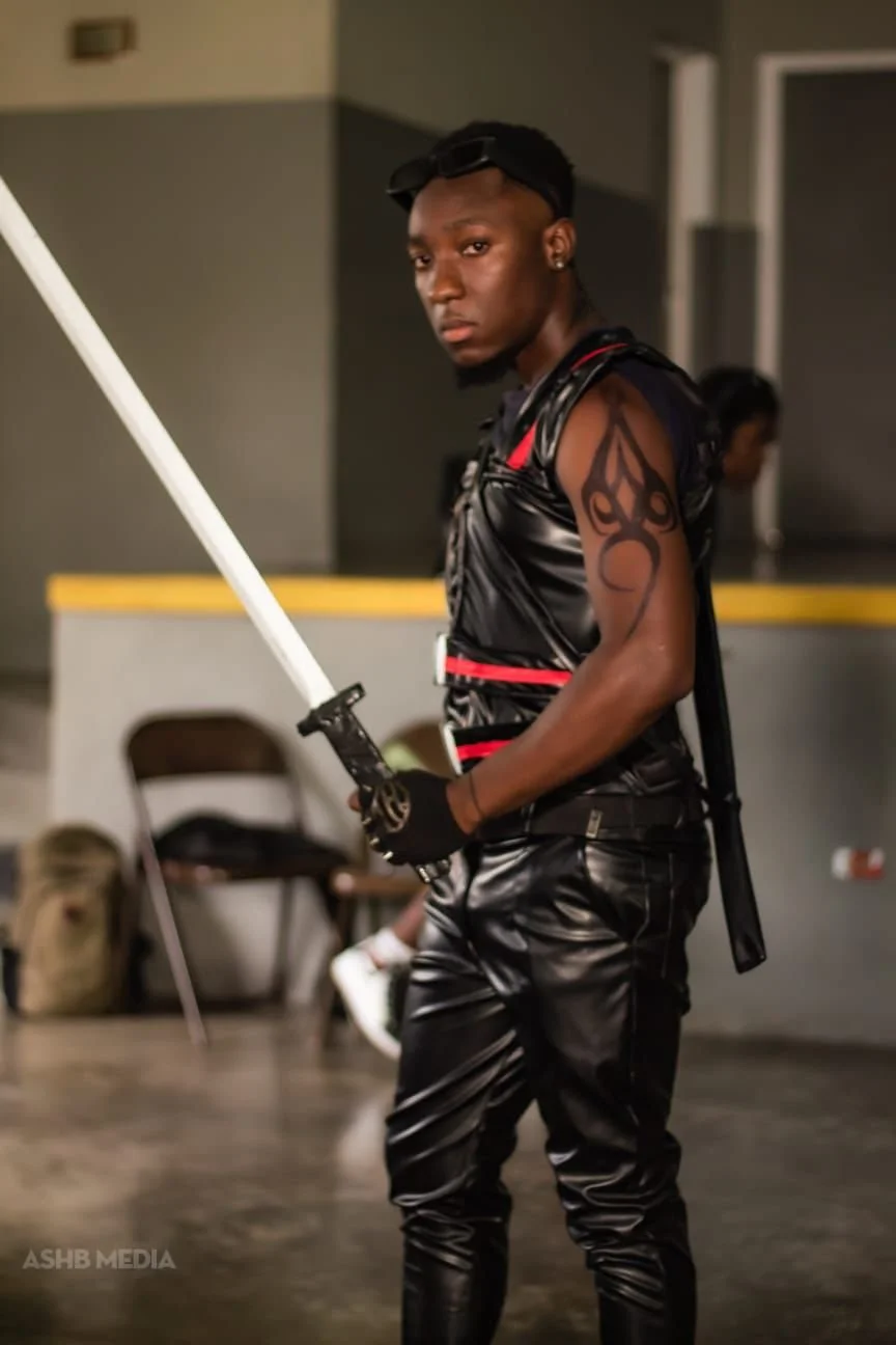 Young man in a black leather outfit holding a sword, standing indoors, with a tattoo on his right arm, wearing sunglasses on his head.