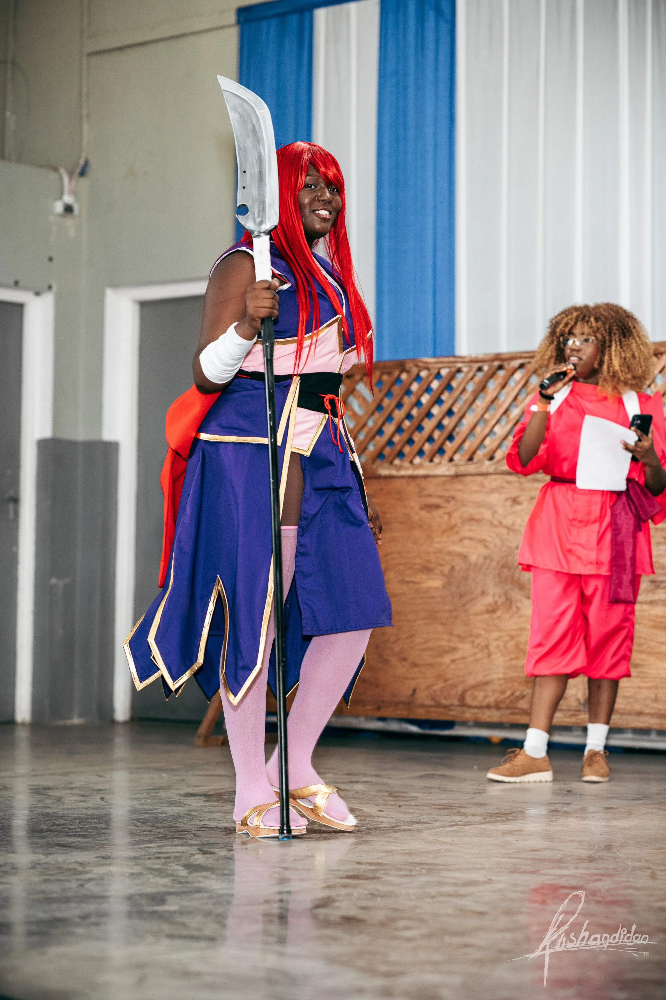 Two women in costumes on stage, one dressed as a warrior with a sword and red wig, the other in a pink outfit with curly hair, holding a microphone and a piece of paper.