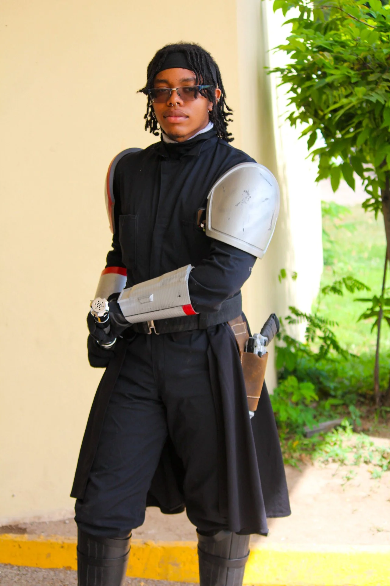 A person dressed in a black outfit with futuristic armor on the shoulders and forearms, wearing sunglasses, standing indoors near a window with green plants outside.