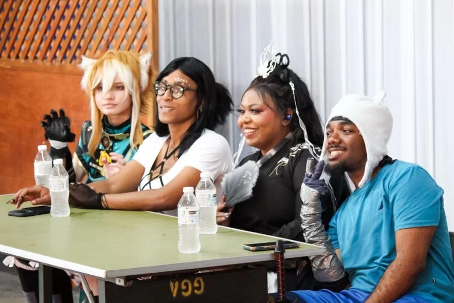 Group of four people seated at a table dressed in cosplay costumes, with water bottles and phones on the table, indoors with metallic and wooden background.