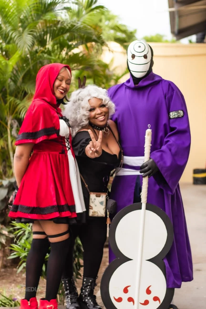 Three people dressed in costumes: a woman in a Little Red Riding Hood dress, a woman with gray curly hair making a peace sign, and a person in a purple robe with a white mask, holding a staff and standing next to a large circular prop. They are outdo