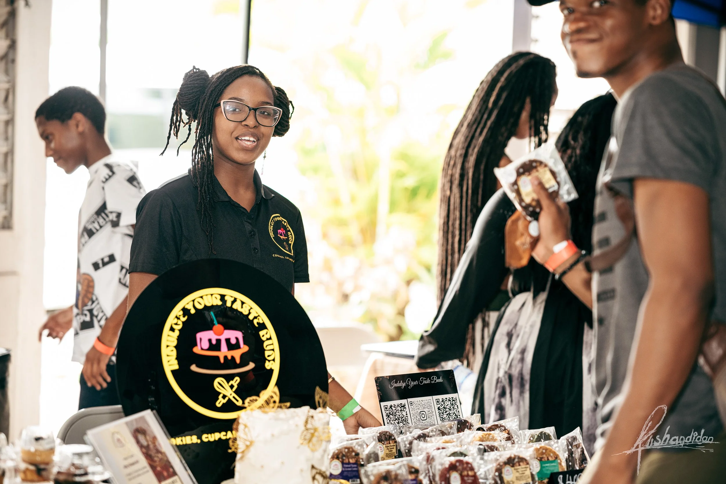 A woman standing behind a table at a food booth with a sign that says "Indulge Your Taste Buds". She is wearing a black shirt with a logo, glasses, and has braided hair. Several food items, including cookies and cupcakes, are displayed on the table. 