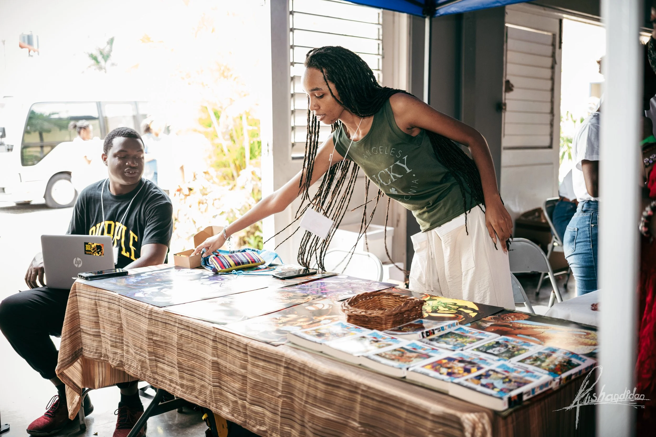 A woman at a table with comics, a small basket, and various items, leaning over to pick something up, while a young man with a laptop watches her.
