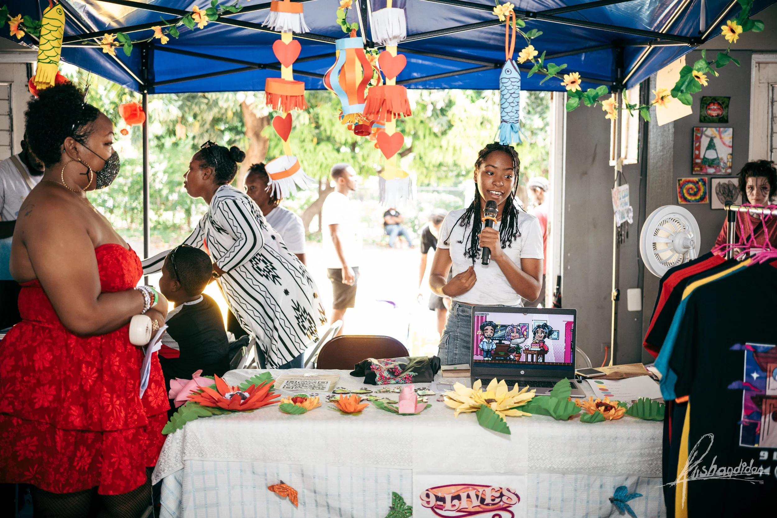 A girl speaking into a microphone at a booth decorated with paper flowers and hanging crafts at a community event. There are people, including women and children, talking and browsing around. The booth has a laptop with cartoon characters displayed o