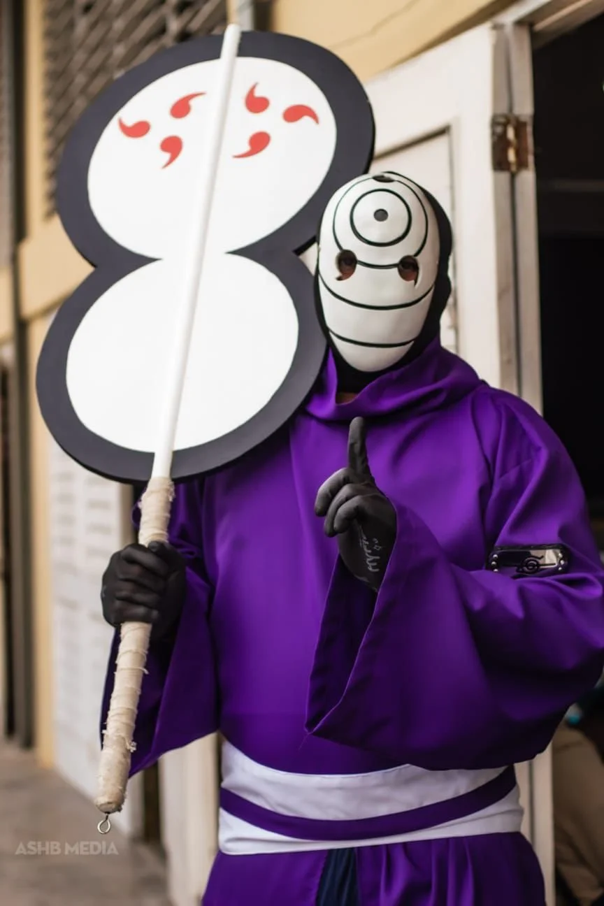 Person dressed as Naruto Uzumaki from Naruto holding a large 8-shaped sign with red symbols, making a shushing gesture with their index finger, wearing a purple costume and a mask with a spiraled pattern.