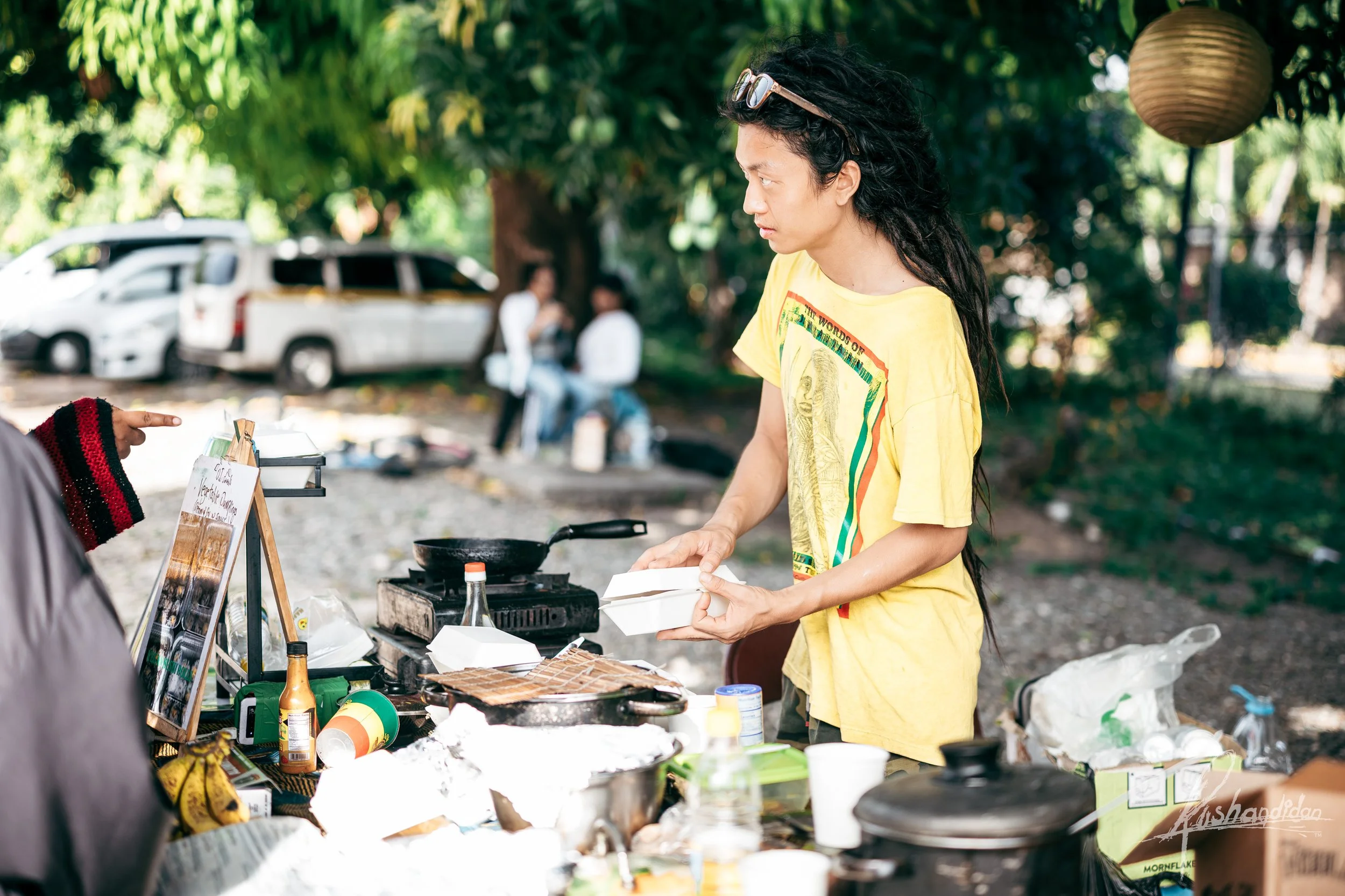 A person in a yellow shirt is preparing food outdoors at a makeshift stall, with various food items, condiments, and cooking utensils on the table. In the background, there are trees, a few parked cars, and two people sitting and talking.