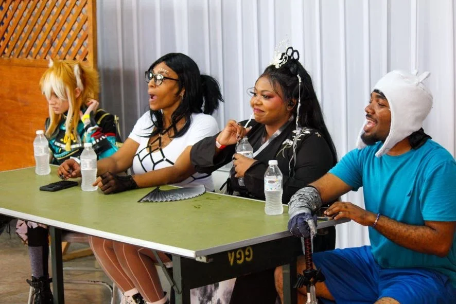 Four individuals sitting at a table dressed in various costumes, with water bottles and a remote control on the table, in a room with a curtain backdrop.