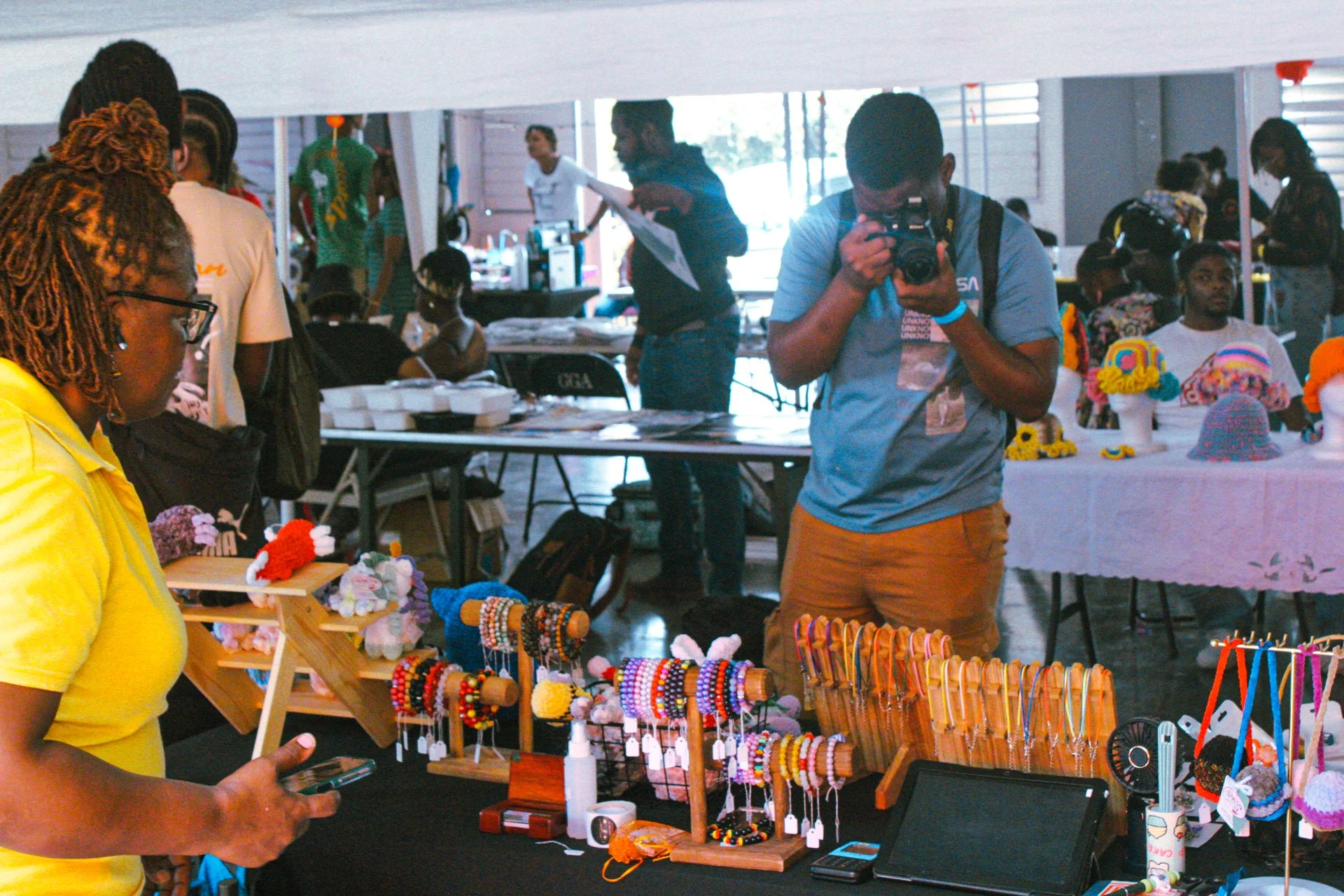 People browsing handmade jewelry and crafts at a market stall, with a woman in yellow examining items and a man taking photos.