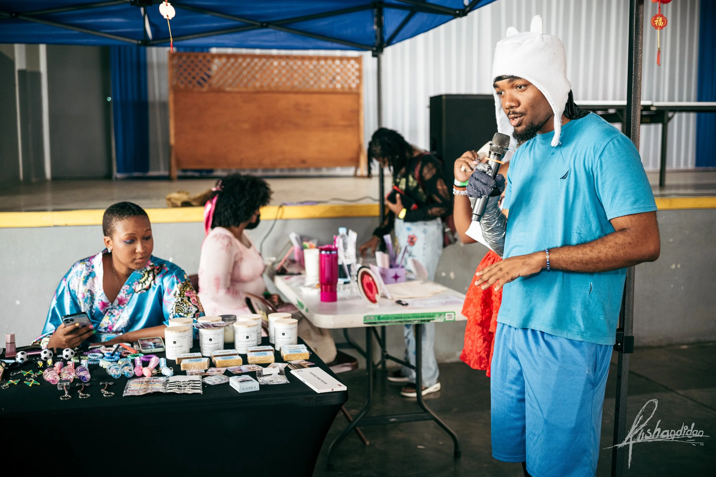 A man in a blue shirt and shorts with a white hat speaking into a microphone at a booth with jewelry and other small items for sale, with women behind the table and on a stage in the background.