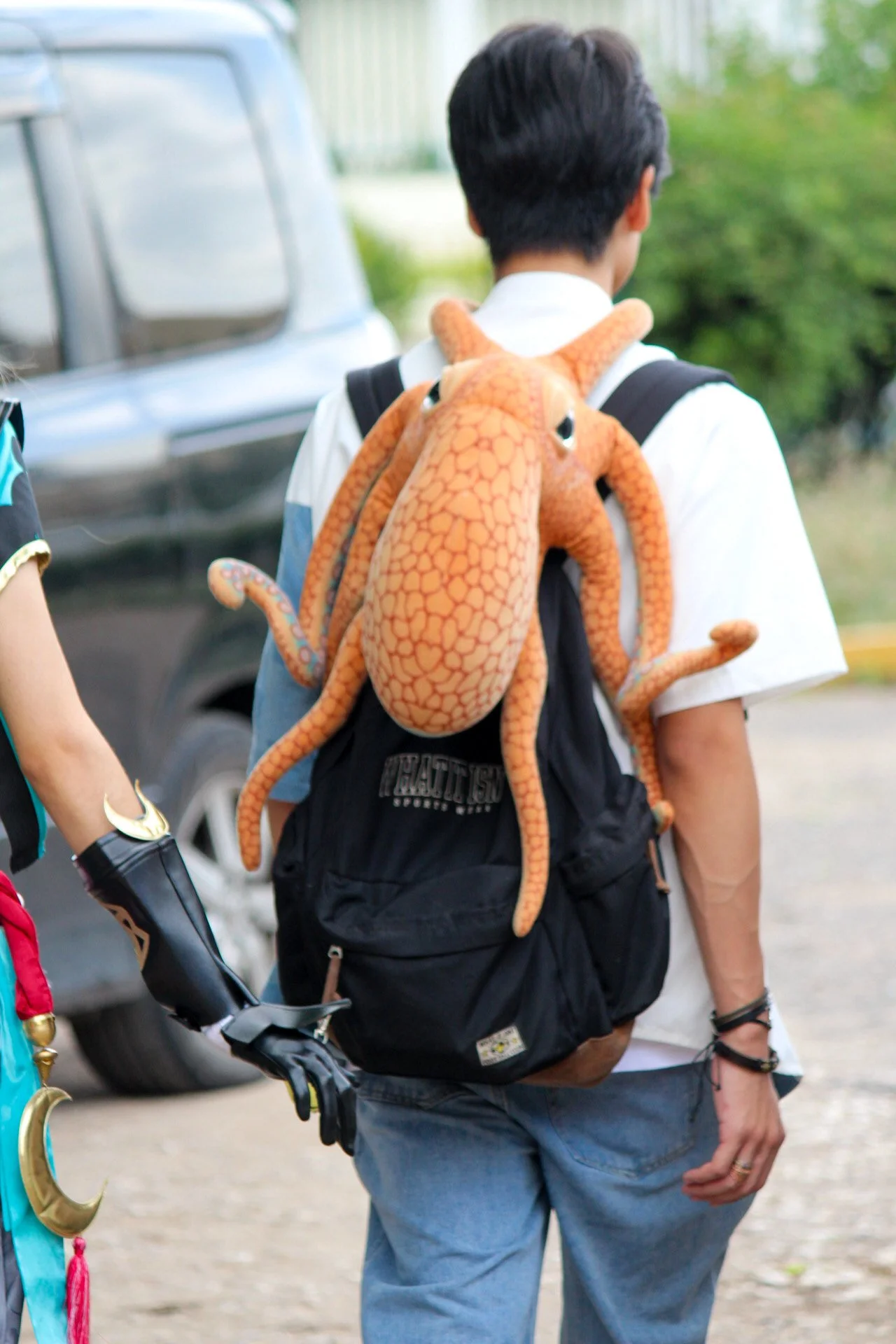A person with dark hair wearing a backpack that has an orange plush octopus tentacle wrapped over their shoulder.
