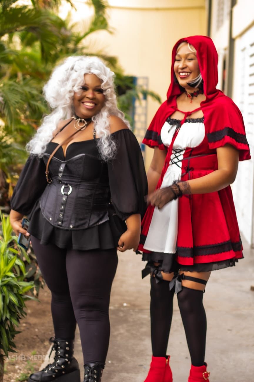 Two women are standing outdoors, both dressed in costumes. The woman on the left is wearing a black witch or gothic style outfit with a corset and a silver wig. The woman on the right is dressed as Little Red Riding Hood with a red cape, red shoes, a