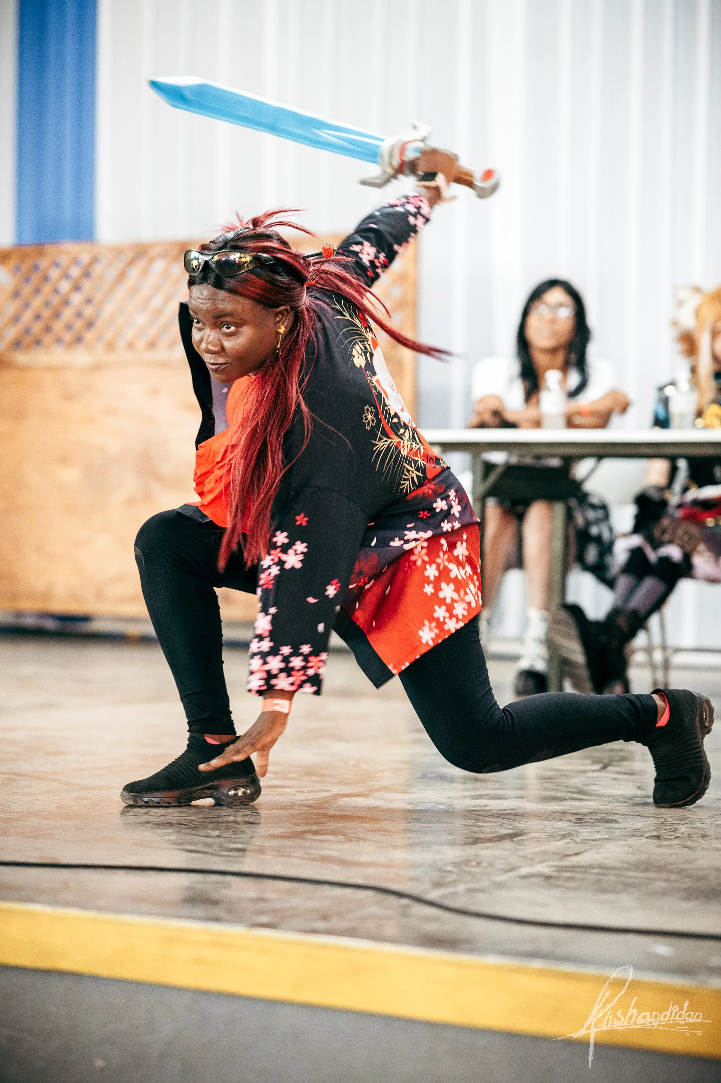 A person dressed as a warrior, holding a sword, is in a dynamic pose on a stage or performance area.