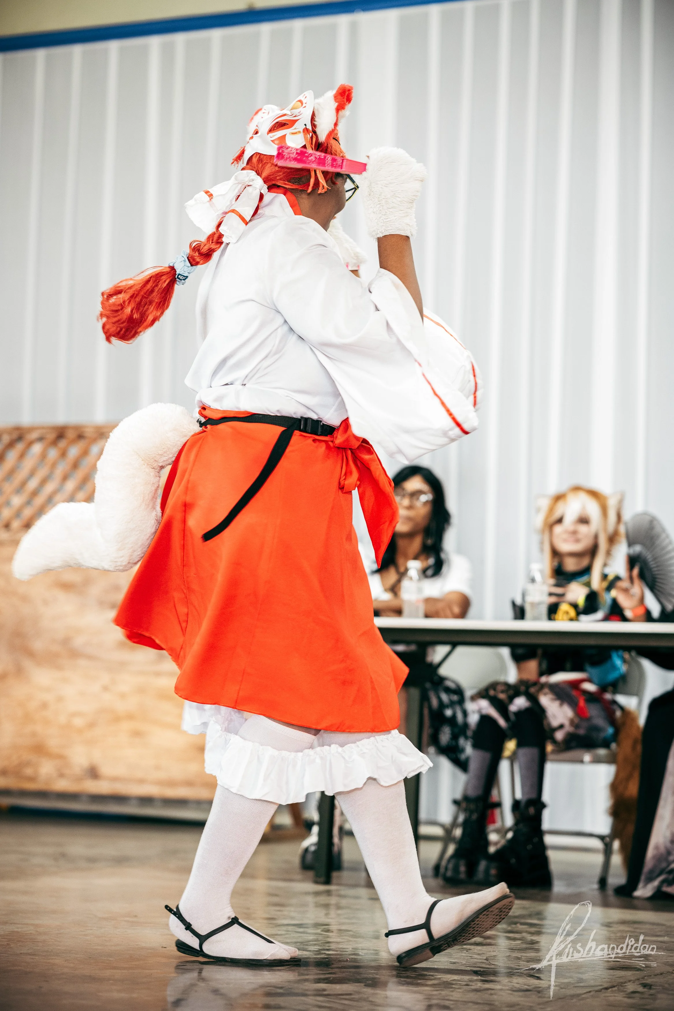Person in a costume with orange skirt, white blouse, white tights, black shoes, and a red wig, holding a white bag, at an indoor event.
