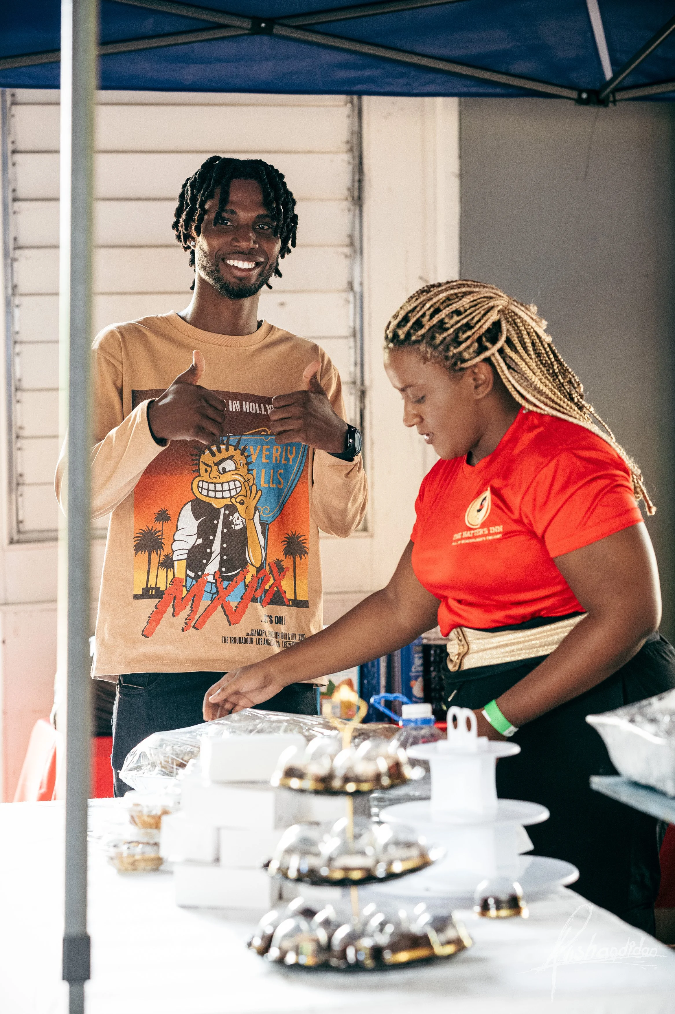 A man smiling and giving a thumbs-up next to a woman working at a table with cakes and desserts at a market stall.