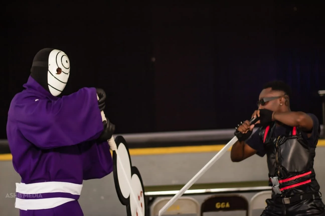 A person in a purple hoodie and mask with a cartoon face design touches a large puppet or mascot, while another person in black sunglasses, face mask, and black gloves holds a white pole, possibly controlling the puppet.