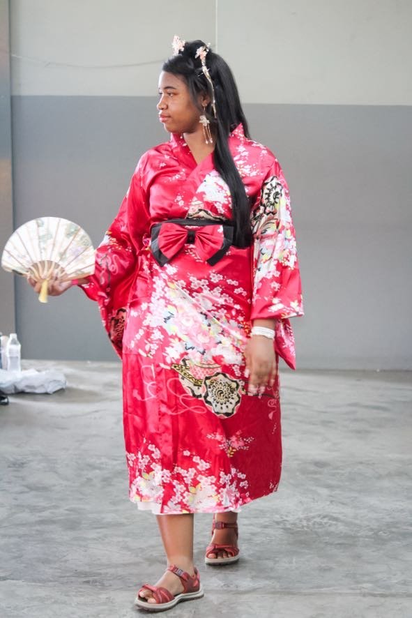 Young woman in a traditional red Japanese kimono with floral and dragon patterns, holding a fan, wearing sandals, standing indoors.