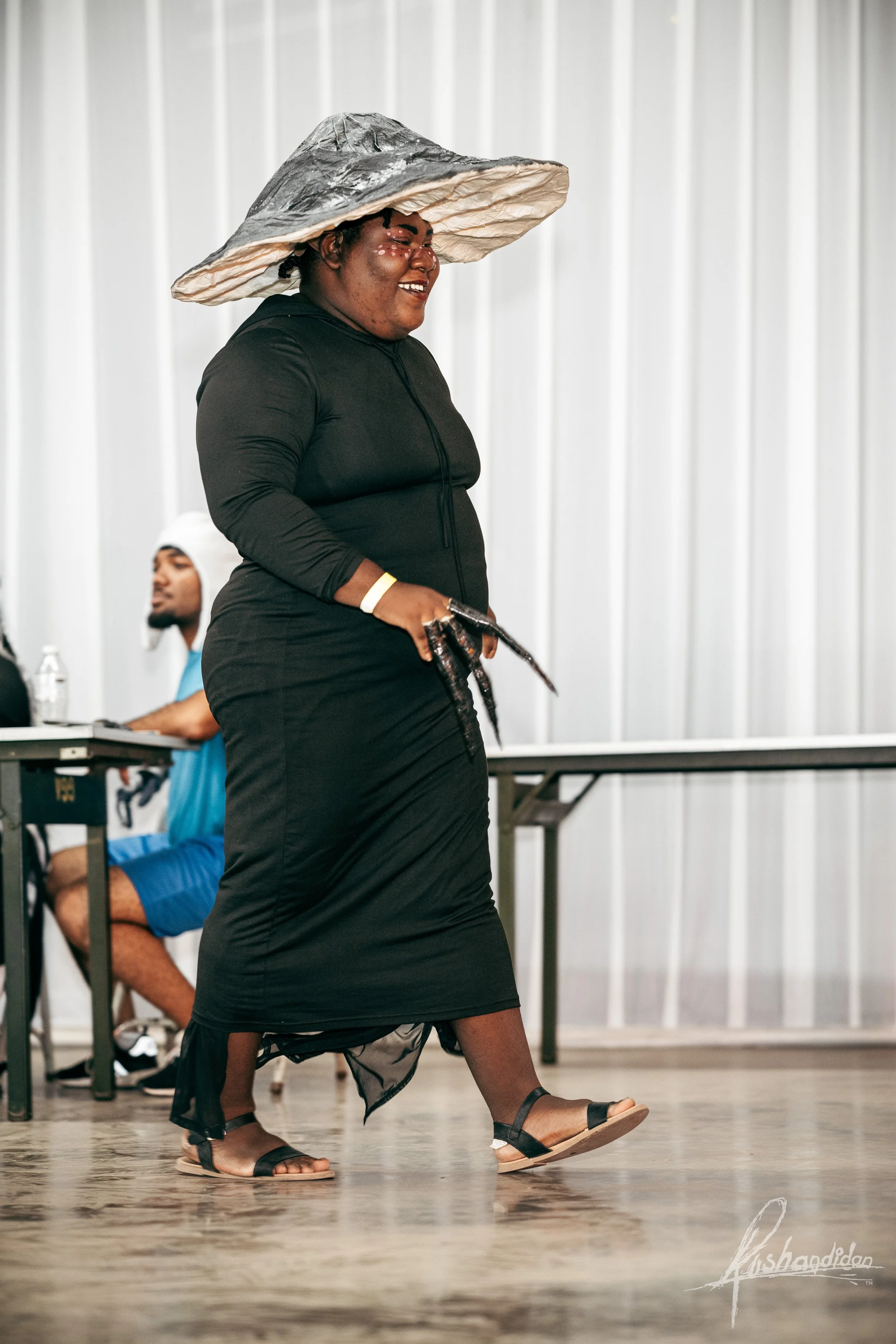 Woman in black dress wearing a large headpiece, smiling, holding objects in her hand, walking indoors with a man sitting at a table in the background.