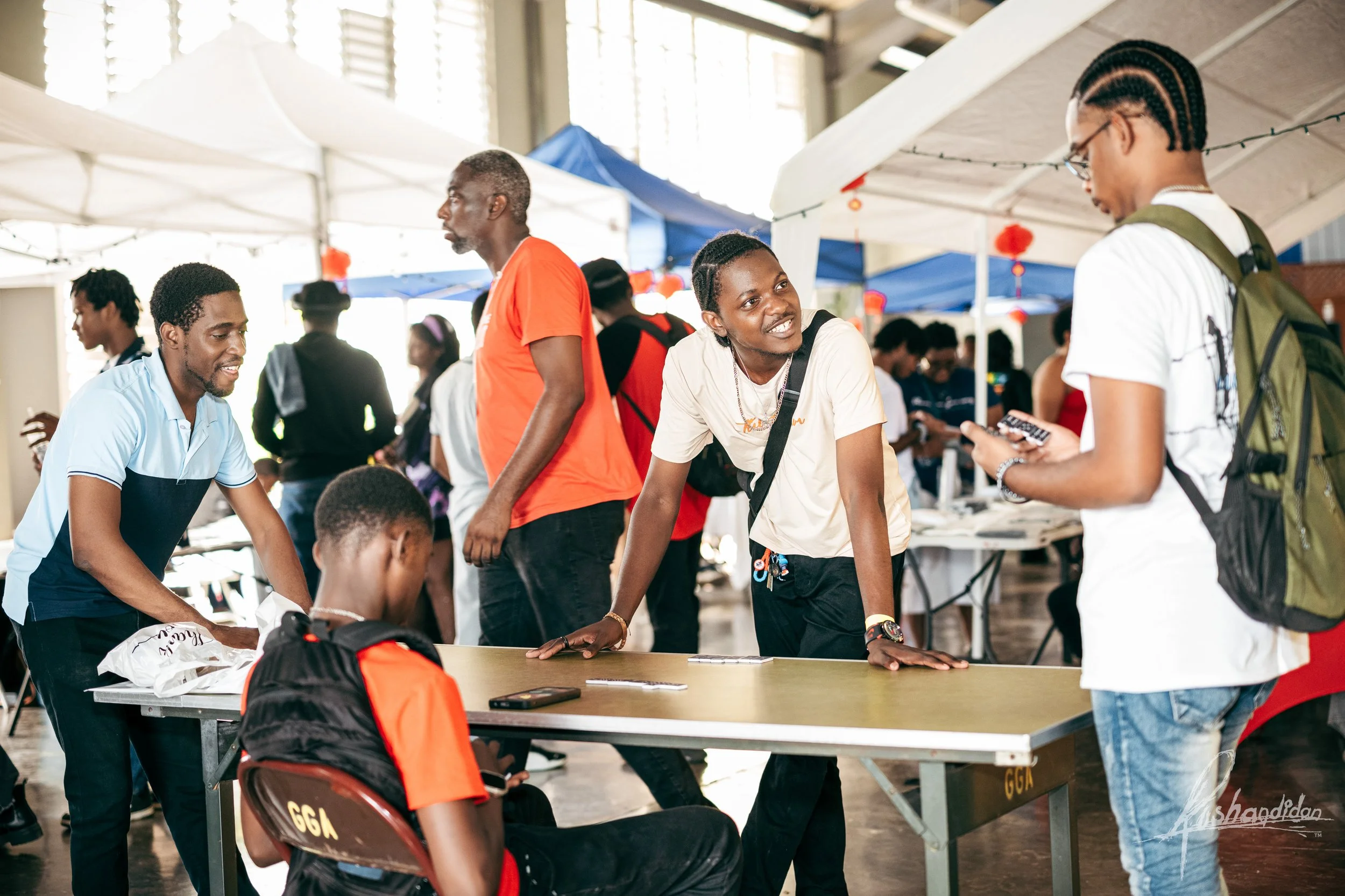Group of young people engaging at a table under white and blue tents at an indoor event, with some individuals smiling and interacting, and others looking at their phones.
