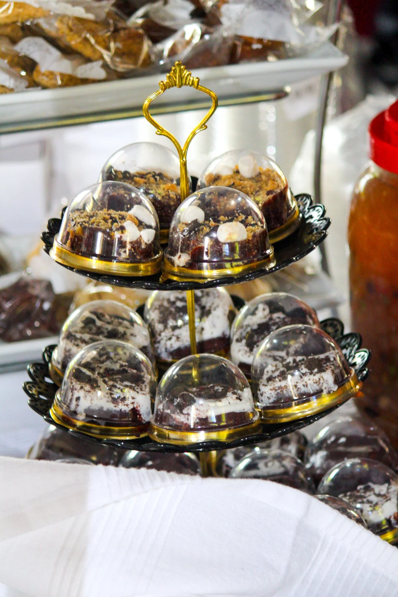 A two-tiered black and gold dessert stand holding small glass domes covering various cakes with white and dark chocolate toppings in a bakery or café setting.